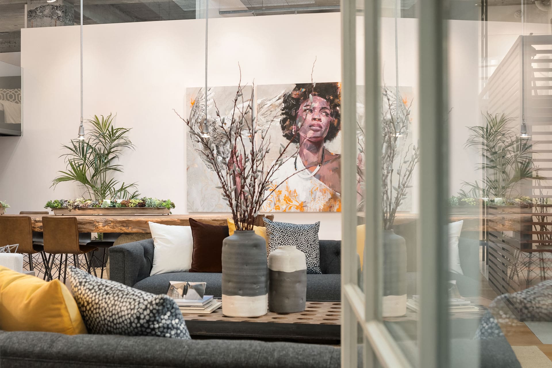 Modern living room with contemporary decor. A large painting of a person is displayed on the wall. The room features a gray couch adorned with various cushions, a wooden coffee table with decorative vases, and plants adding a touch of greenery.