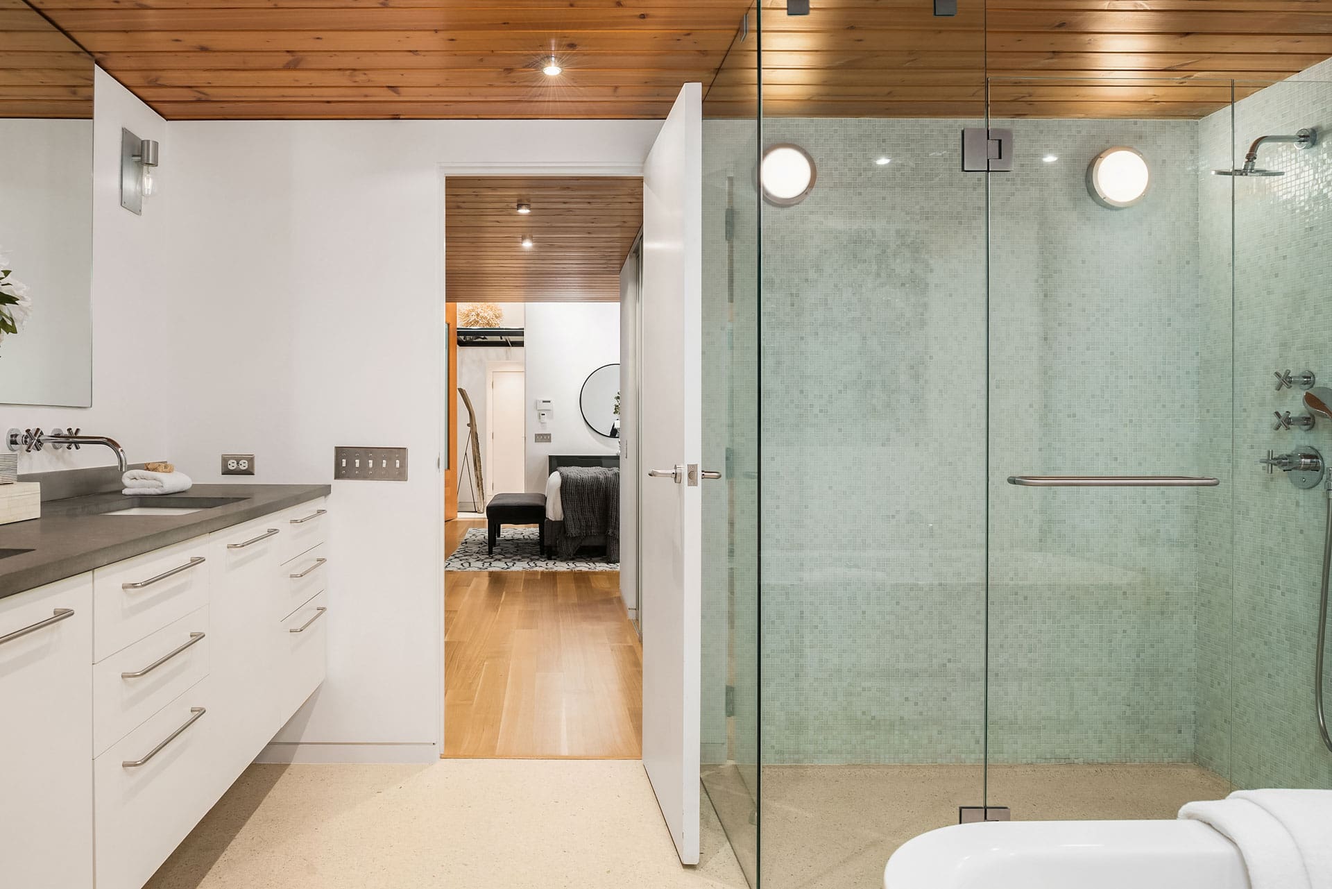 Modern bathroom with a glass-enclosed shower, light green tiled walls, and a wooden ceiling. The bathroom includes a white vanity with a gray countertop and a mirror. Through an open door, a bedroom with wooden floor can be seen in the background.