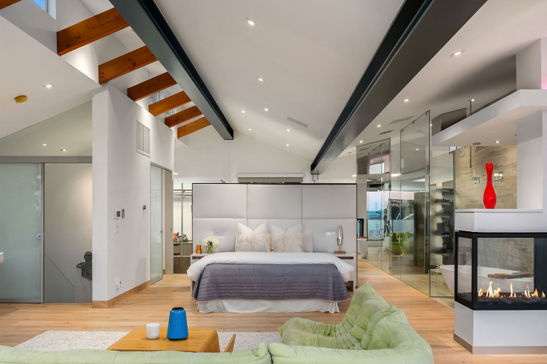 A modern bedroom with a high, beamed ceiling and wooden floors features a cozy bed with white and gray bedding. A glass-enclosed fireplace serves as a partition between the sleeping area and the living space, which includes a green sofa and a blue vase on a coffee table.