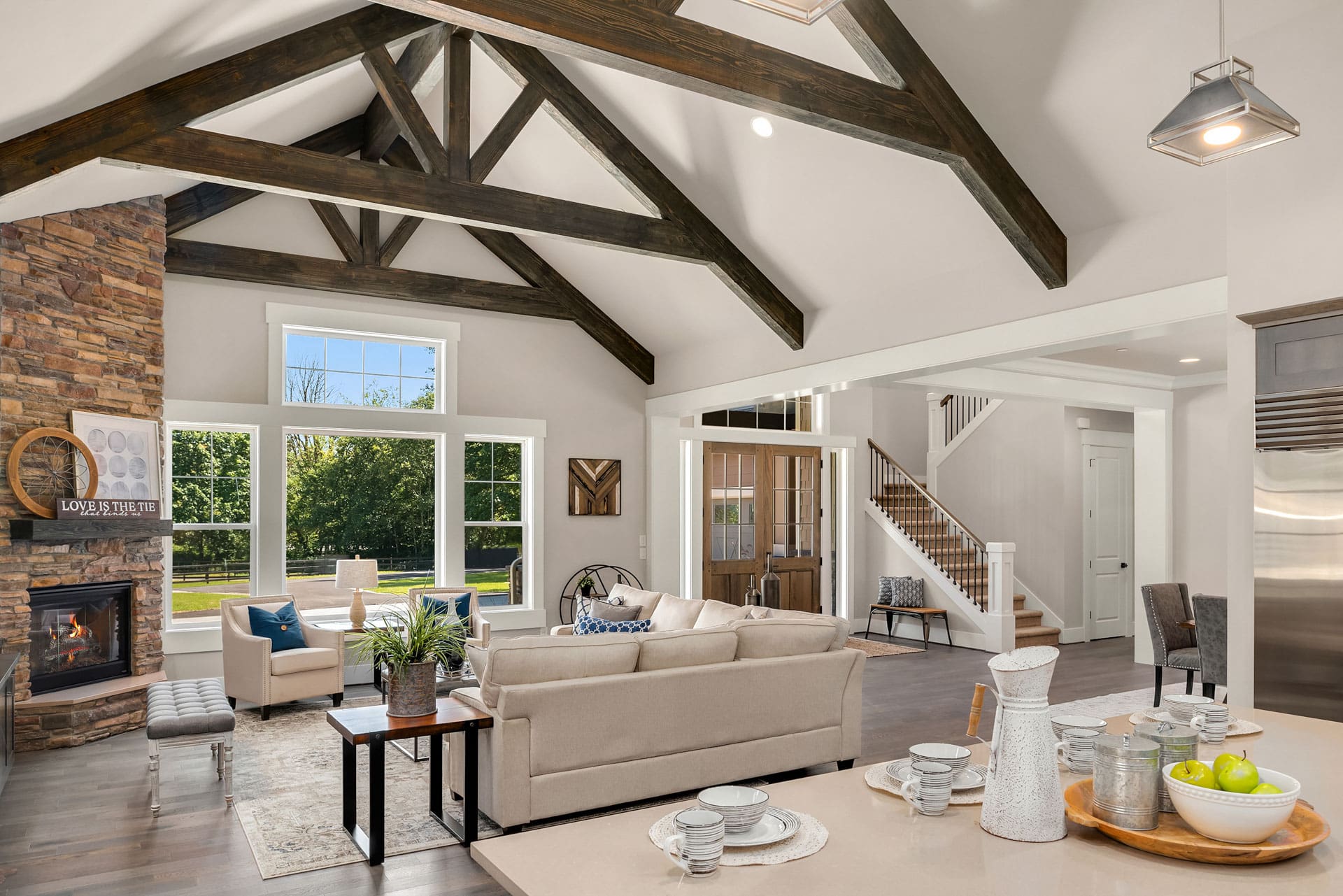 A spacious, open-concept living room with vaulted ceilings and exposed wooden beams. The room features a stone fireplace, large windows with garden views, and modern, neutral-colored furniture. A dining area and staircase are visible in the background.