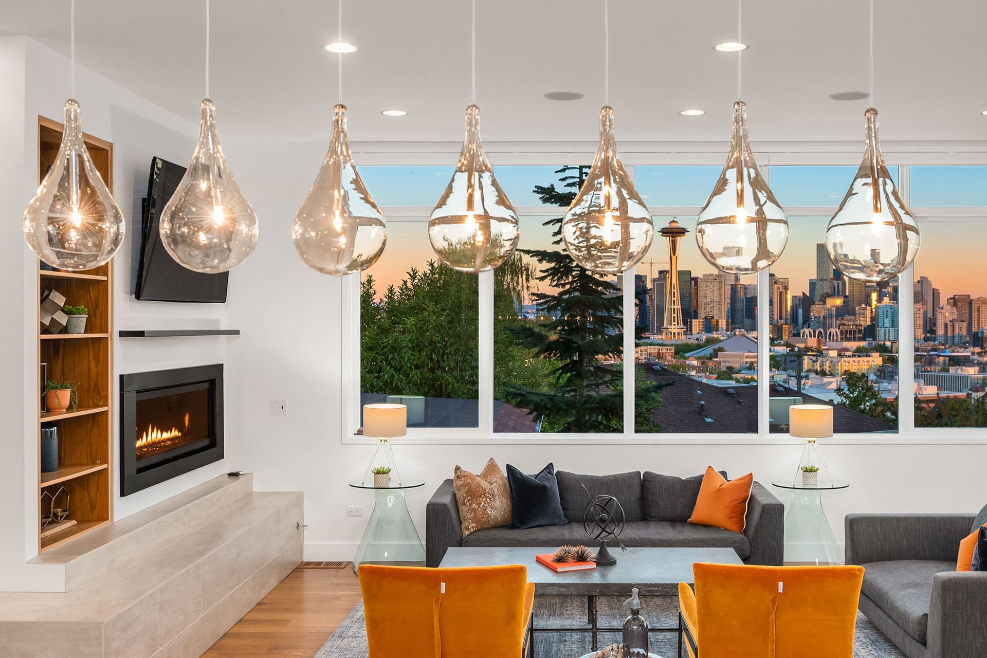 A modern living room with a large window view of a city skyline at sunset. The room features hanging glass light fixtures, a lit fireplace, gray sofas, orange chairs, a glass coffee table, and built-in wooden shelves. Large windows showcase a scenic cityscape.