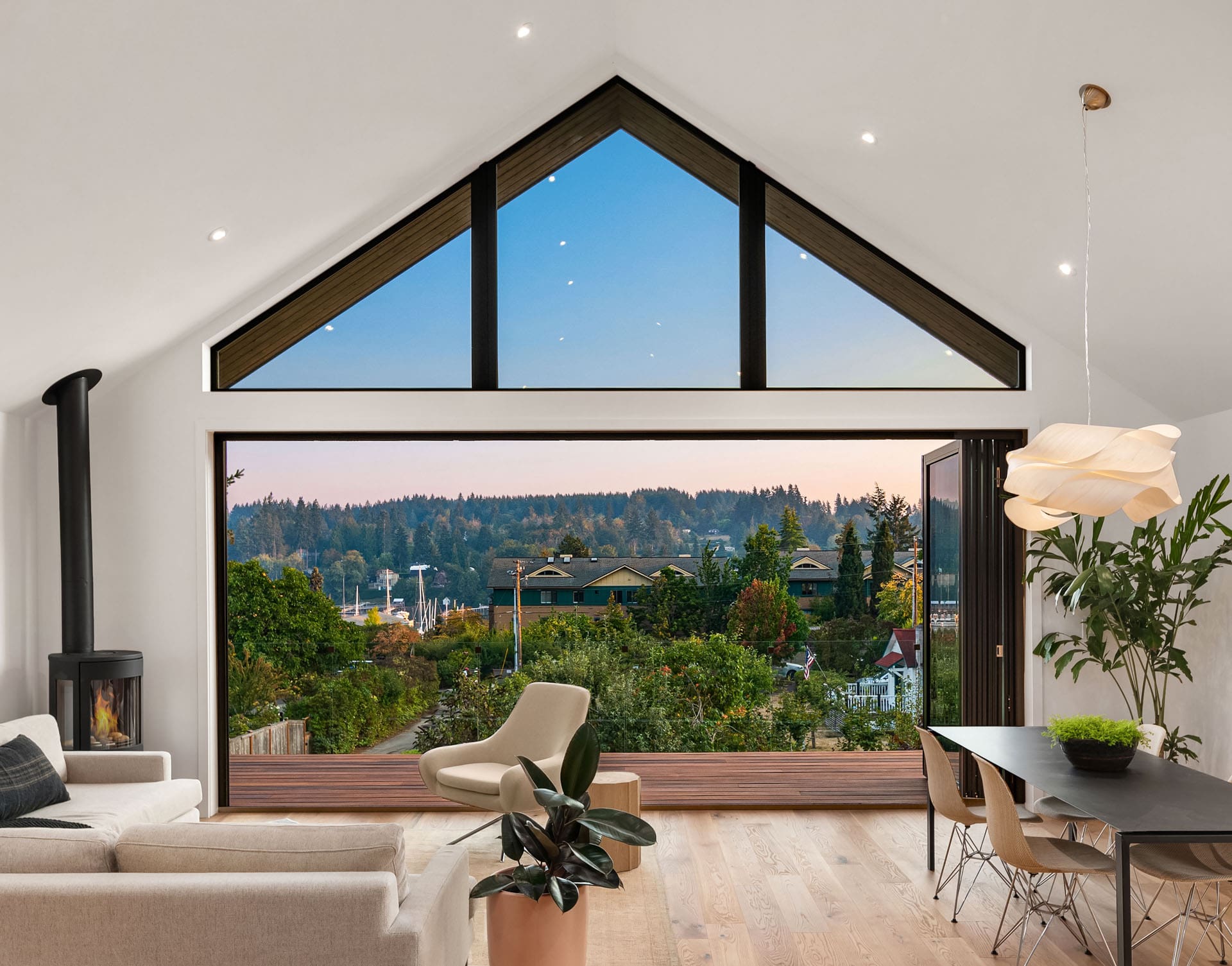 Modern living space with large triangular window offering scenic view of forested hills. Room features light furniture, round wood stove, potted plants, dining table with chairs, and warm wooden flooring. High ceiling with recessed lighting enhances brightness.