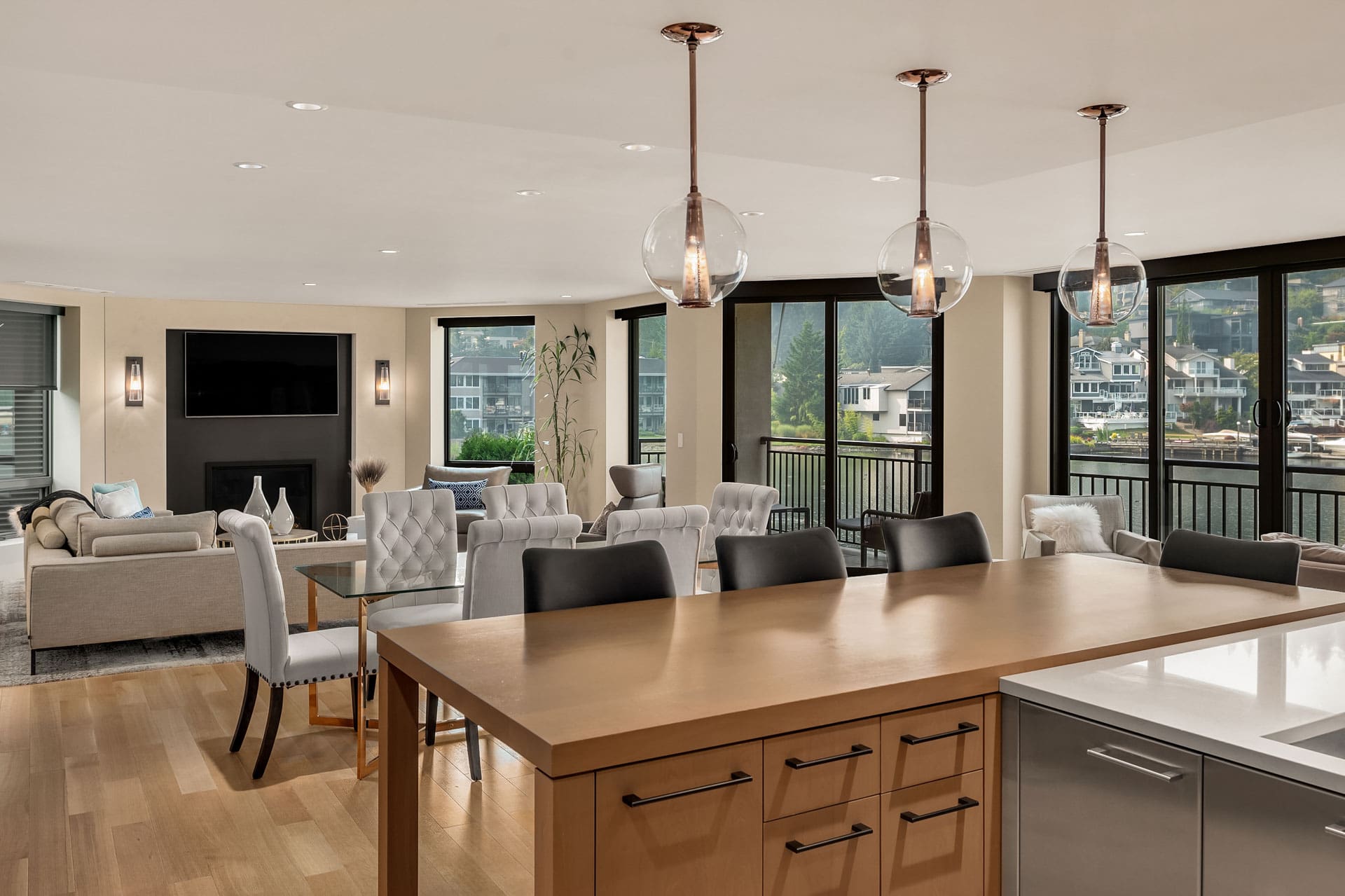 A modern open-plan living space with hardwood floors, featuring a kitchen island with pendant lights, a dining table surrounded by tufted chairs, and a cozy seating area with a couch and coffee table. Floor-to-ceiling windows offer views of houses across a body of water.