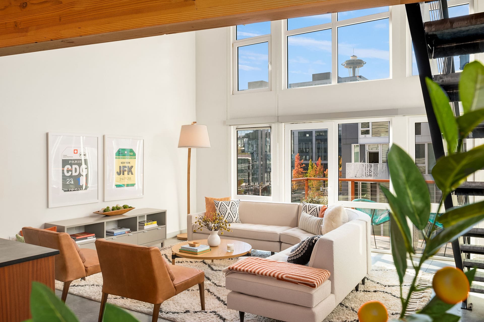 A spacious, sunlit living room featuring a beige sectional sofa with assorted pillows, two orange armchairs, a tall floor lamp, and a low bookshelf. Large windows provide a view of a cityscape, while indoor plants add a touch of greenery to the modern decor.