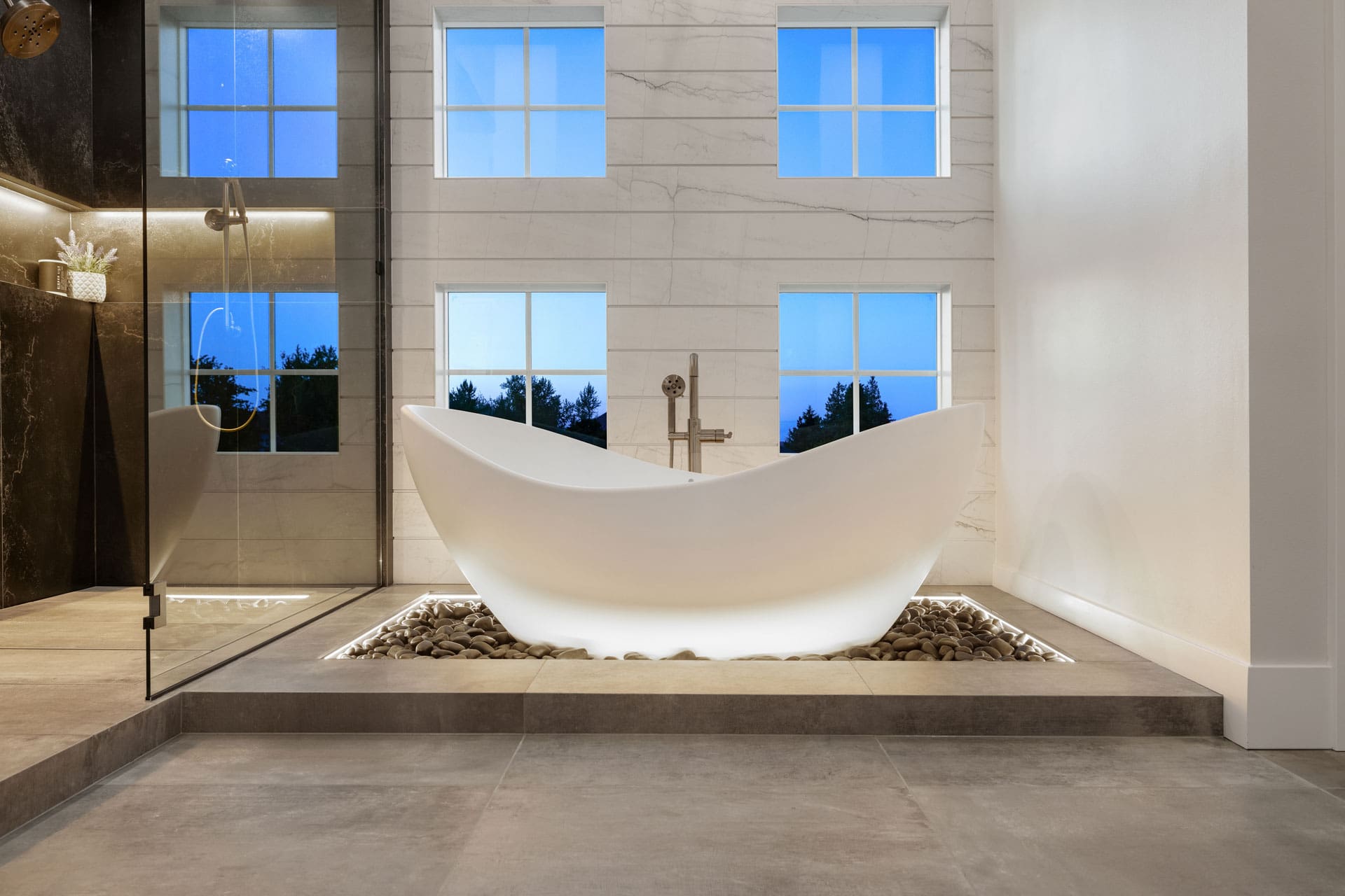 Modern bathroom featuring a sleek white freestanding bathtub on a bed of smooth stones with recessed lighting. The room has large windows showcasing a twilight sky, a glass-enclosed shower with black tiles, and minimalistic decor for a serene ambiance.