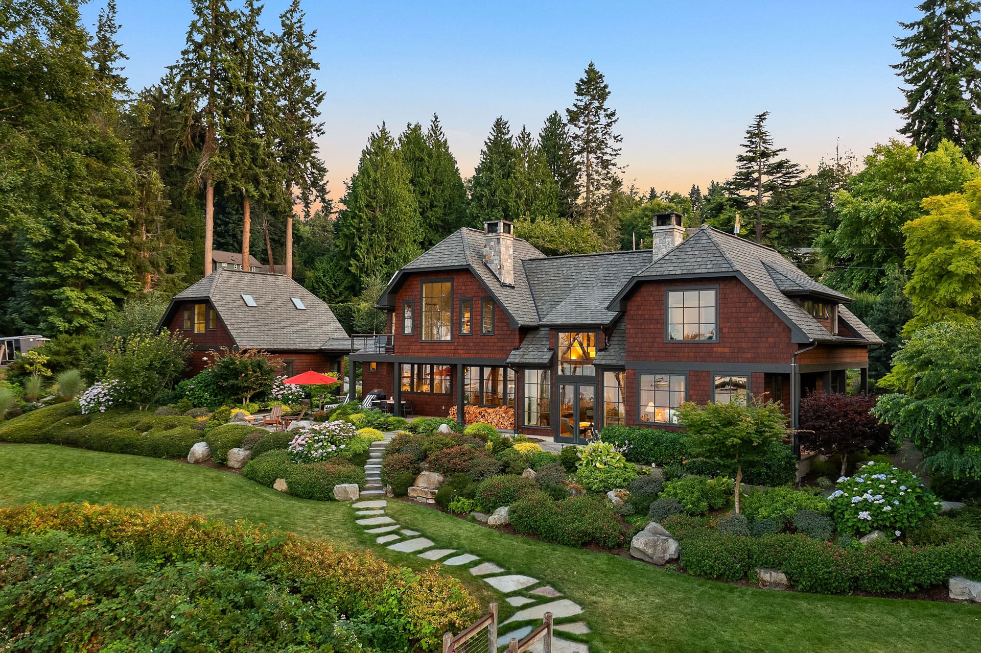 A large, rustic house with dark brown exterior and multiple windows sits surrounded by lush greenery and tall trees. A manicured lawn with a stone pathway leads to the entrance, and several shrubs and flowers are scattered throughout the landscape in the forefront.