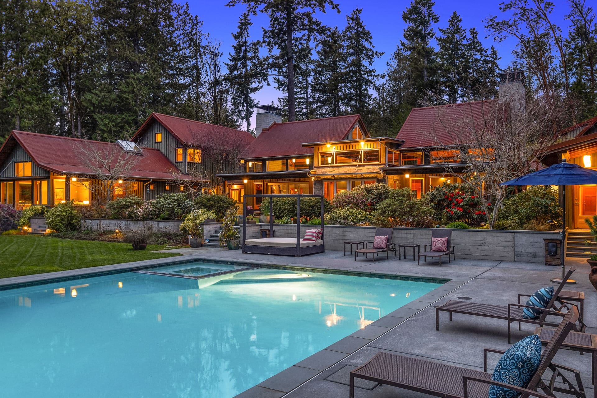 A luxurious, expansive home with red roofs is surrounded by lush greenery and tall trees. The property features extensive outdoor living spaces, including a large, illuminated swimming pool, lounge chairs, and a cozy sitting area under a pergola. The sky is twilight blue.