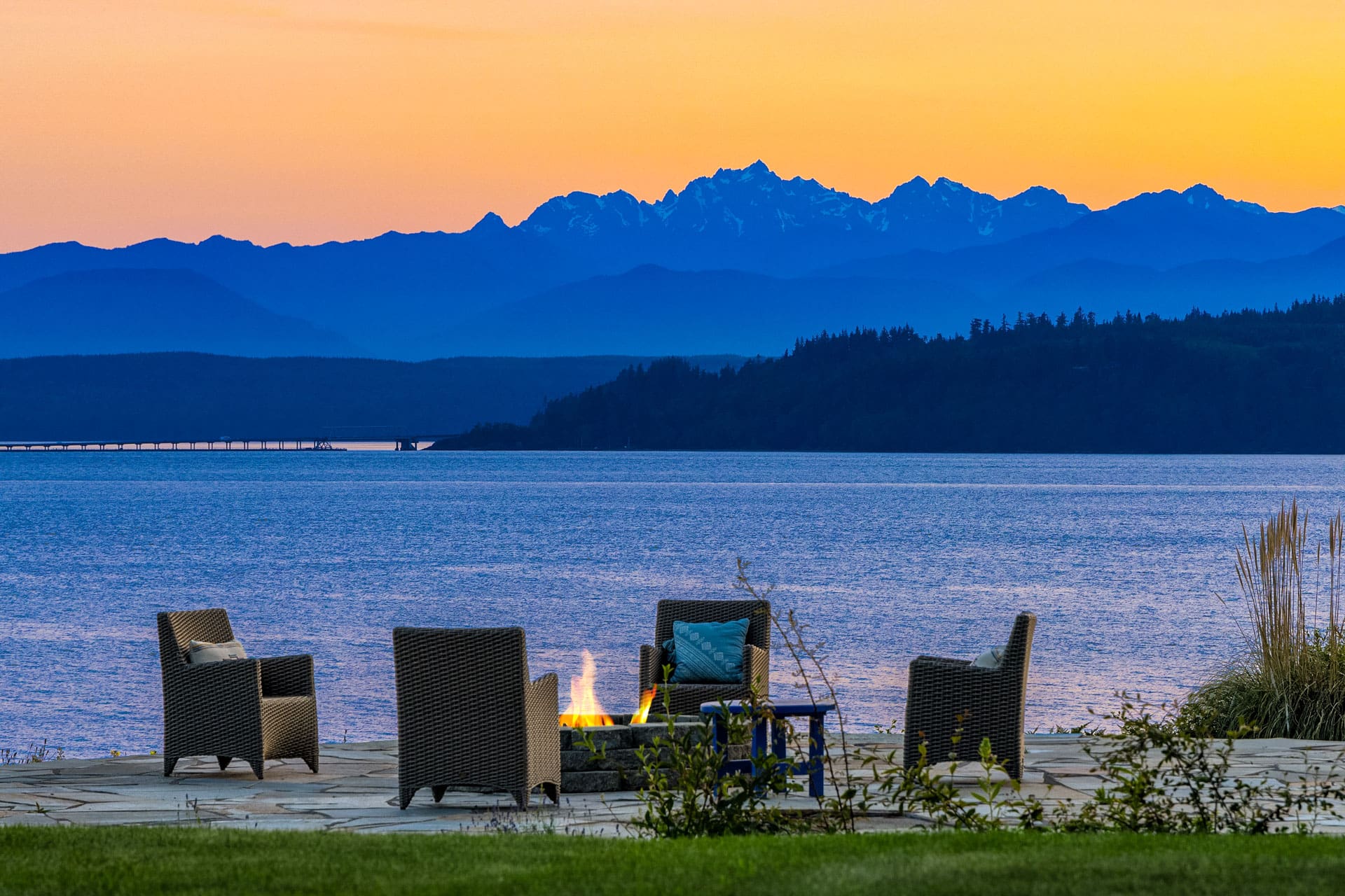 This image shows a lakeside seating area with four wicker chairs around a lit fire pit at sunset. The serene water reflects the orange and blue hues of the sky, and the scene is framed by silhouetted mountains in the distance.