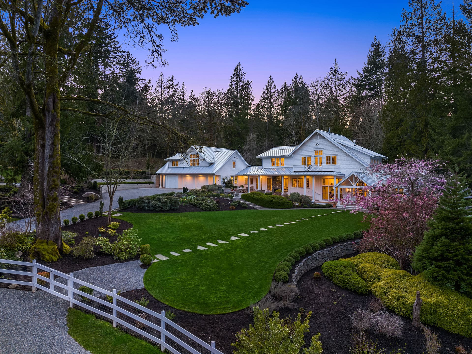 A sprawling white house with a pitched roof sits amid lush greenery framed by trees at twilight. A well-maintained lawn with a stepping stone path leads to the entrance. Flowering bushes and a white fence adorn the landscaped garden.