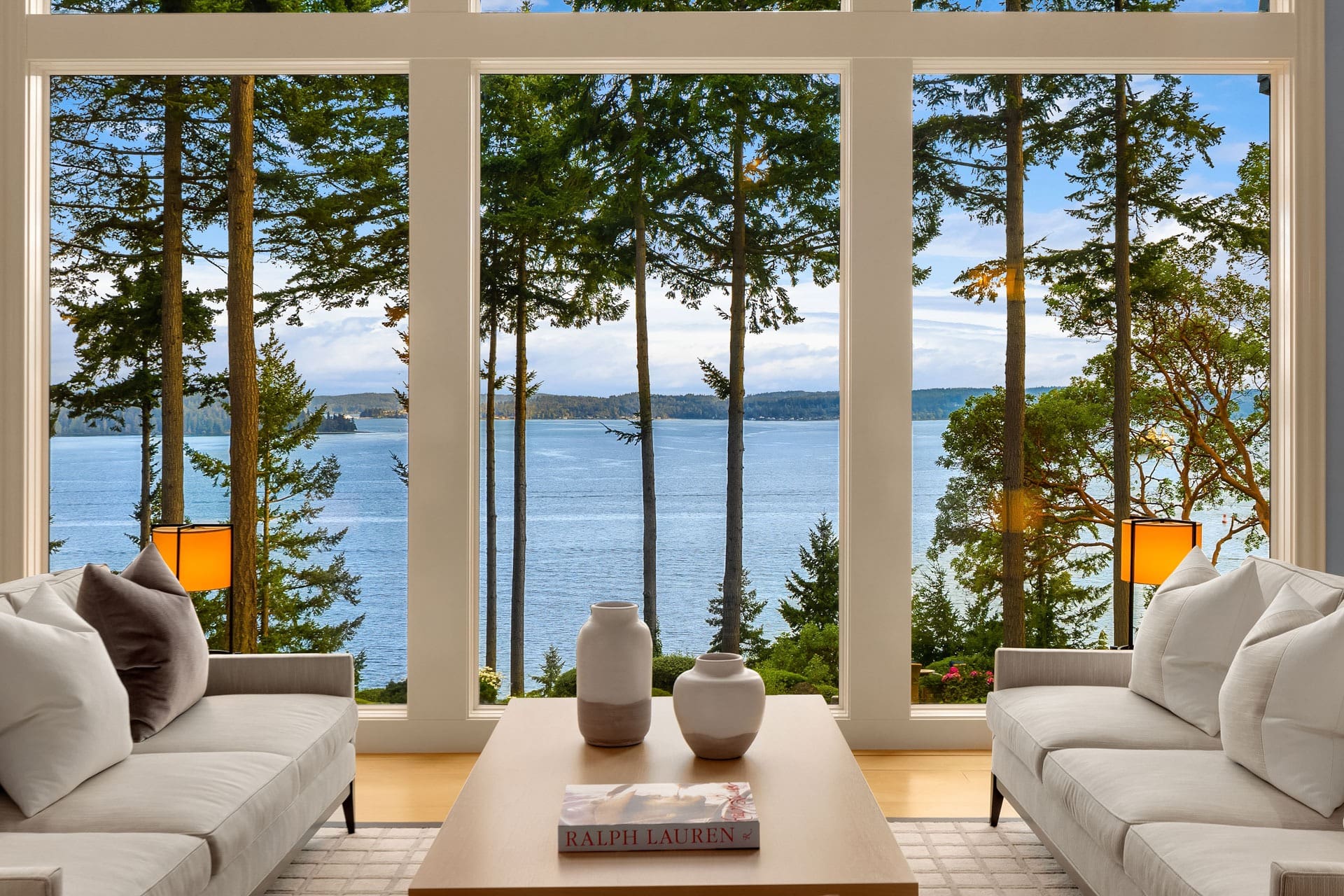 A spacious living room with large floor-to-ceiling windows offering a stunning view of a serene lake and tall trees. The room features two white sofas facing each other, a wooden coffee table with decorative vases, and a book titled "Ralph Lauren.