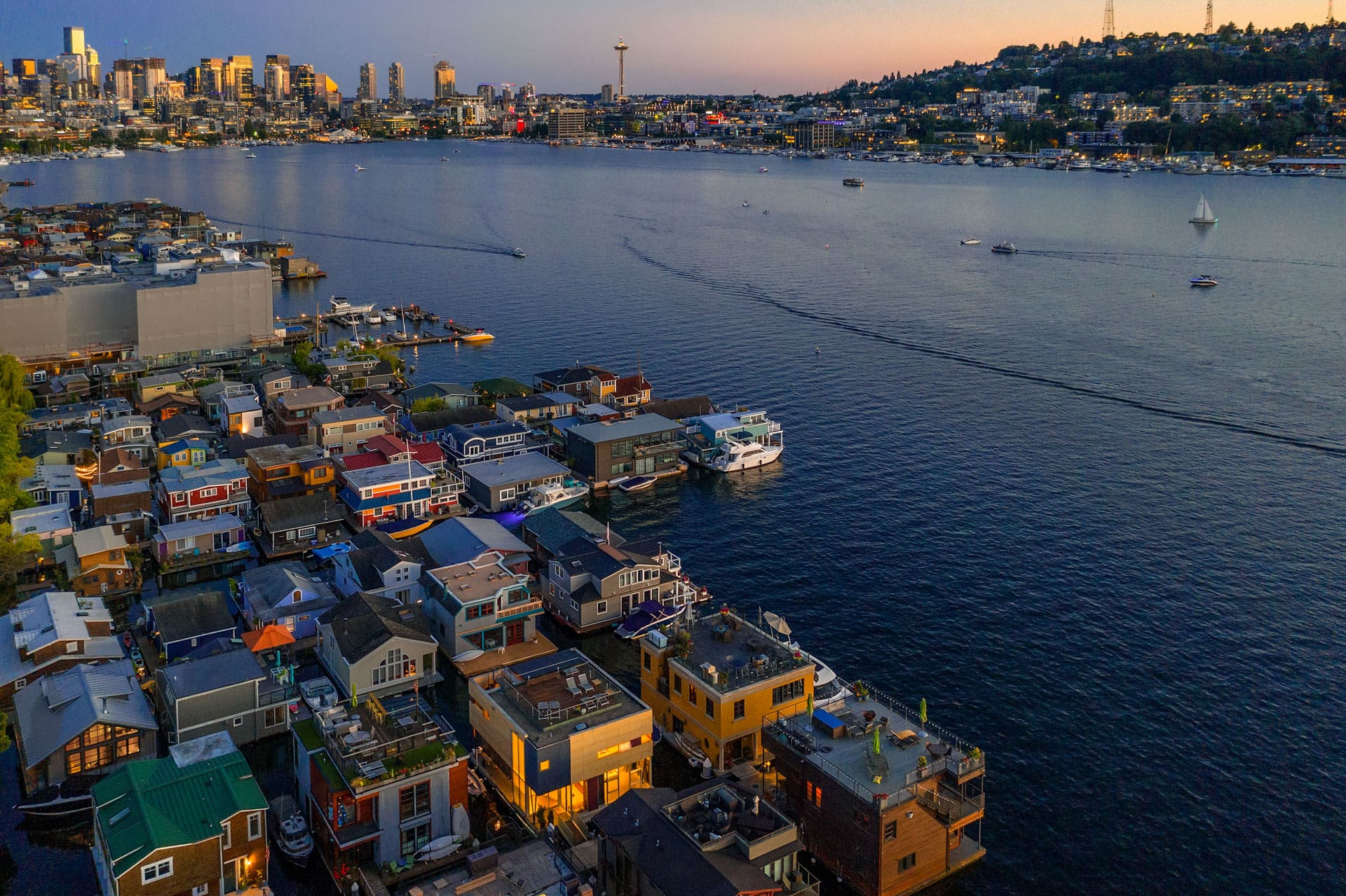 Aerial view of a lake with numerous houseboats in the foreground and a city skyline in the background, including the prominent Space Needle. Several boats are visible on the water during dusk, and city lights are starting to illuminate the scene.