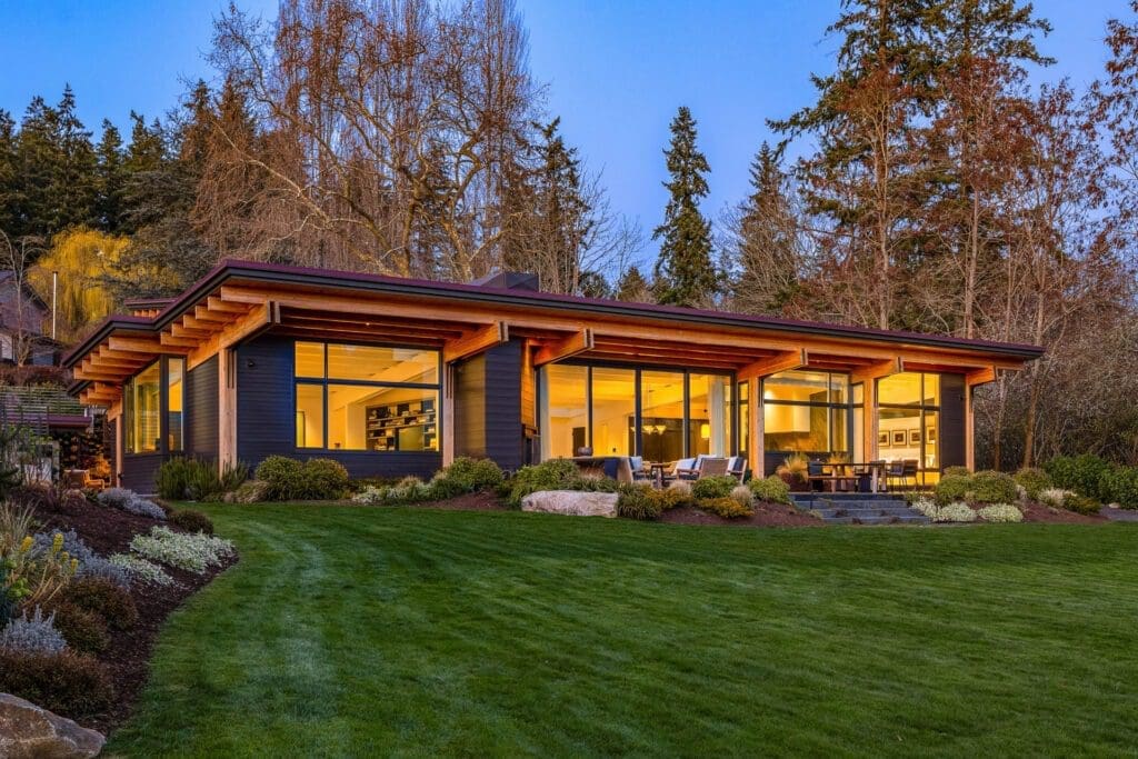 Modern single-story house with large glass windows, wooden exterior, and pitched roof, illuminated warmly from the inside. The house is surrounded by a well-maintained lawn, trees, and a patio with outdoor seating, situated in a serene, forested area during twilight.