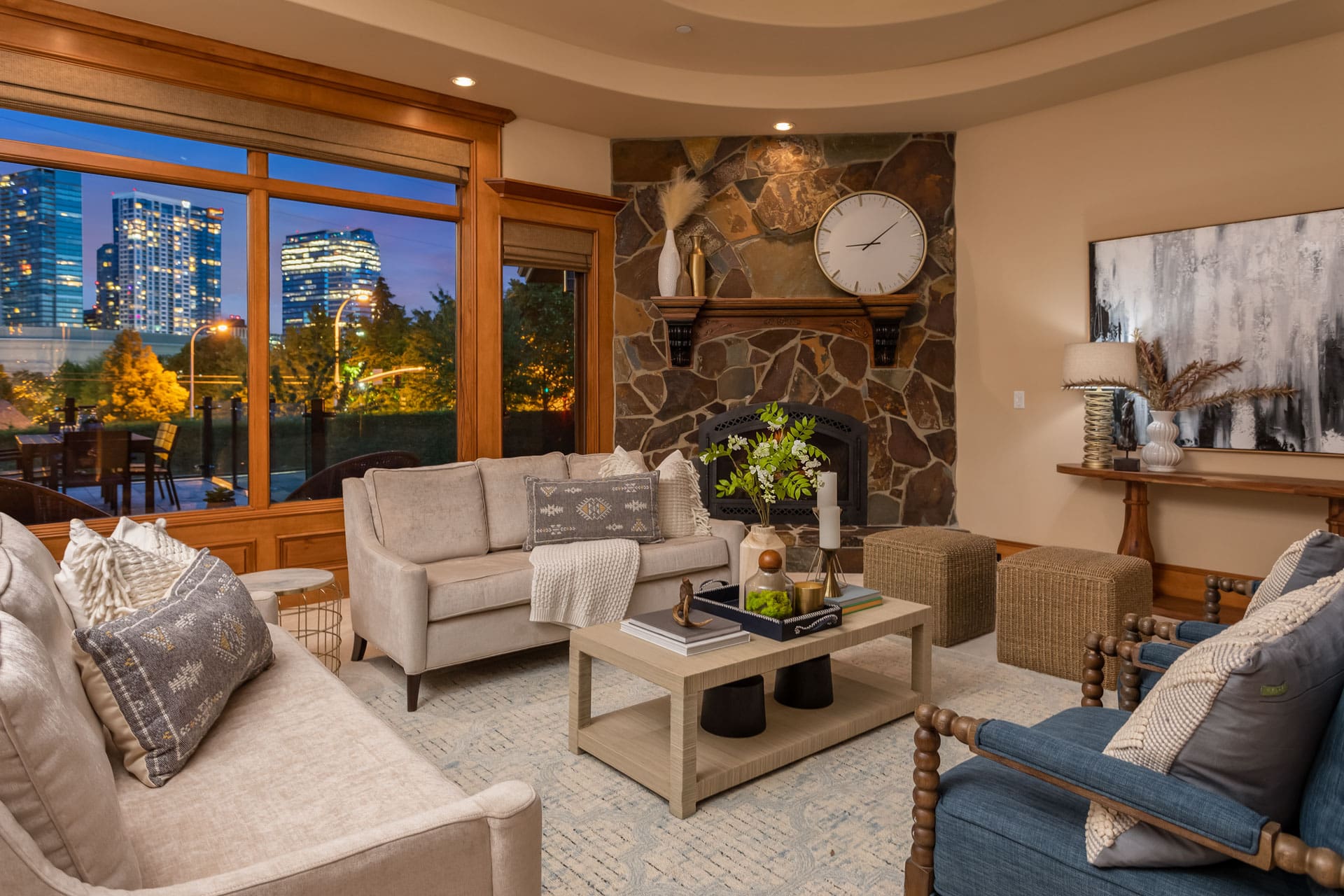 A cozy living room with beige sofas, a stone fireplace, and a large clock above the mantel. There's a coffee table with decorative items and a large abstract painting on the wall. The window offers a view of city buildings lit up at dusk.