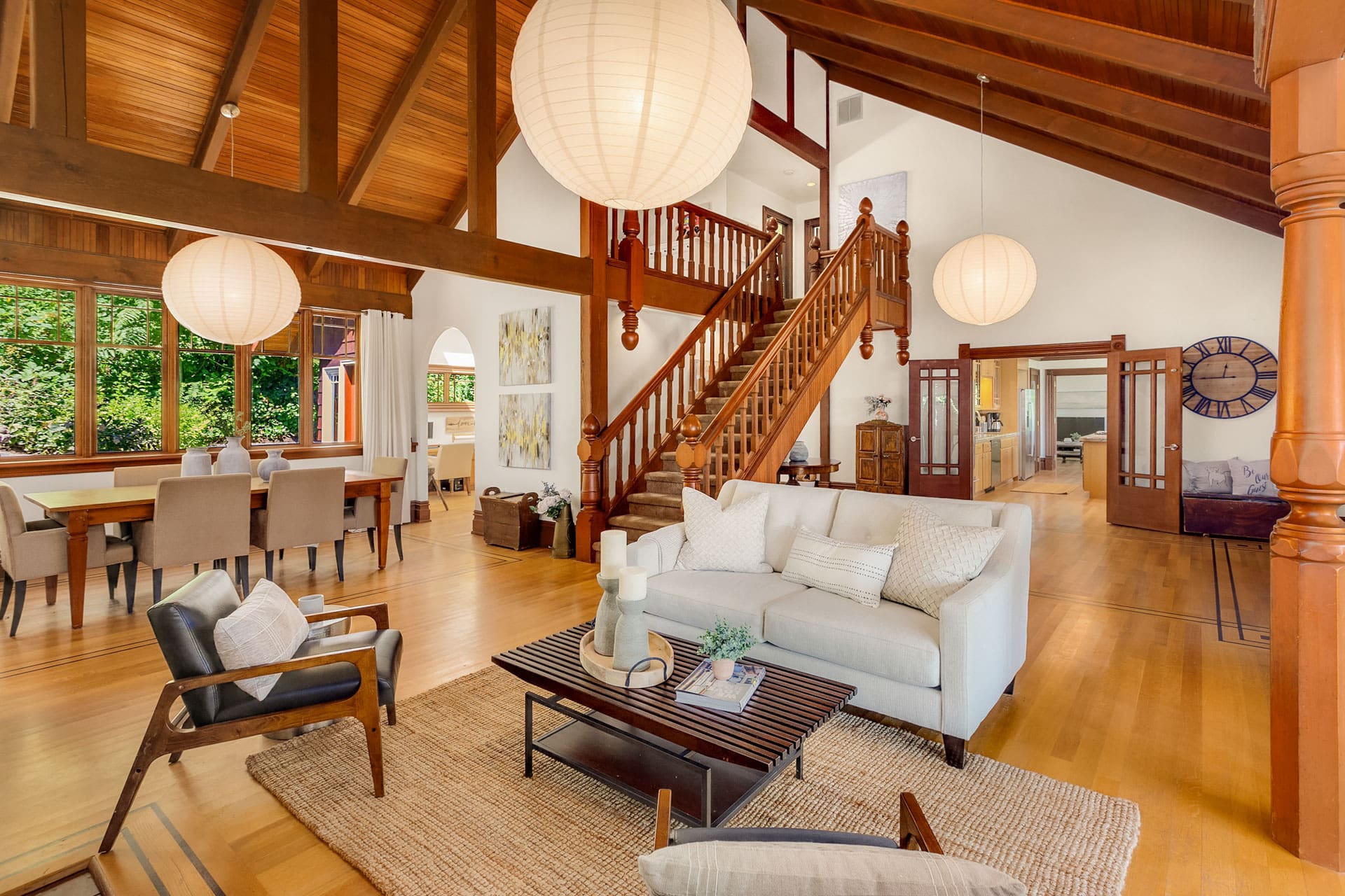 A spacious, well-lit living area with wooden vaulted ceilings and large, spherical pendant lights. It features a comfortable seating arrangement, a coffee table, and a rug. A staircase with wooden railings leads to the upper level. The dining area is visible in the background.