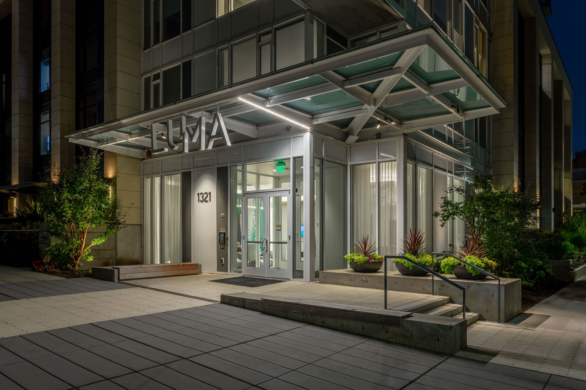 Night view of the entrance to a modern building labeled "LUMA" with "1321" above the door. The entrance is well-lit, with glass doors and large windows. Planters with greenery and a bench are positioned outside. The structure features a geometric awning.