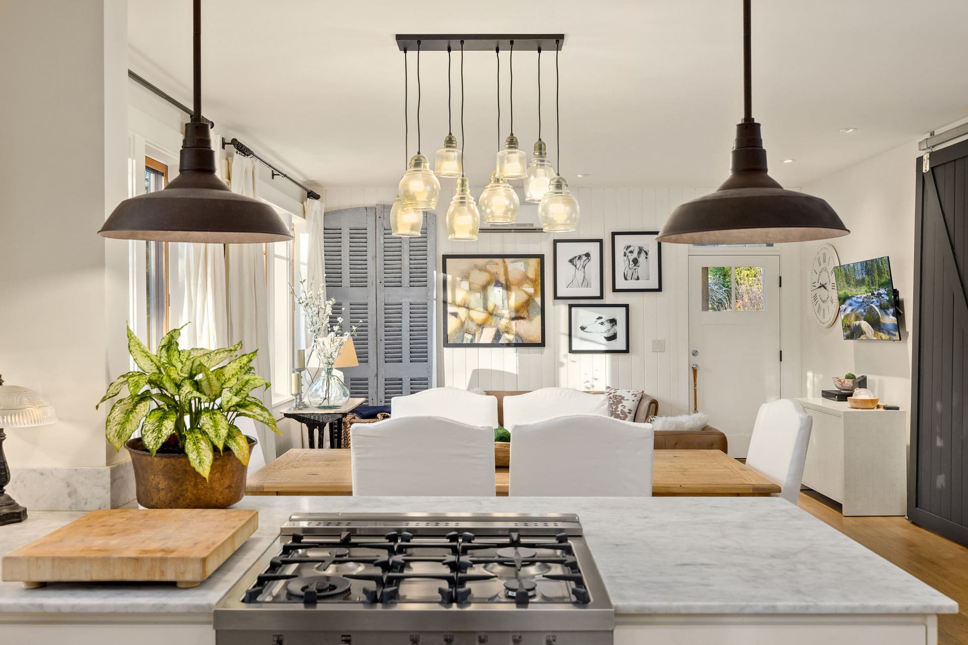 Modern kitchen with a marble island equipped with a gas stove and cutting board. Above the island hangs an array of pendant lights. Behind the island is a dining area with a wooden table and white chairs. Walls display various framed art. Curtains adorn the windows.