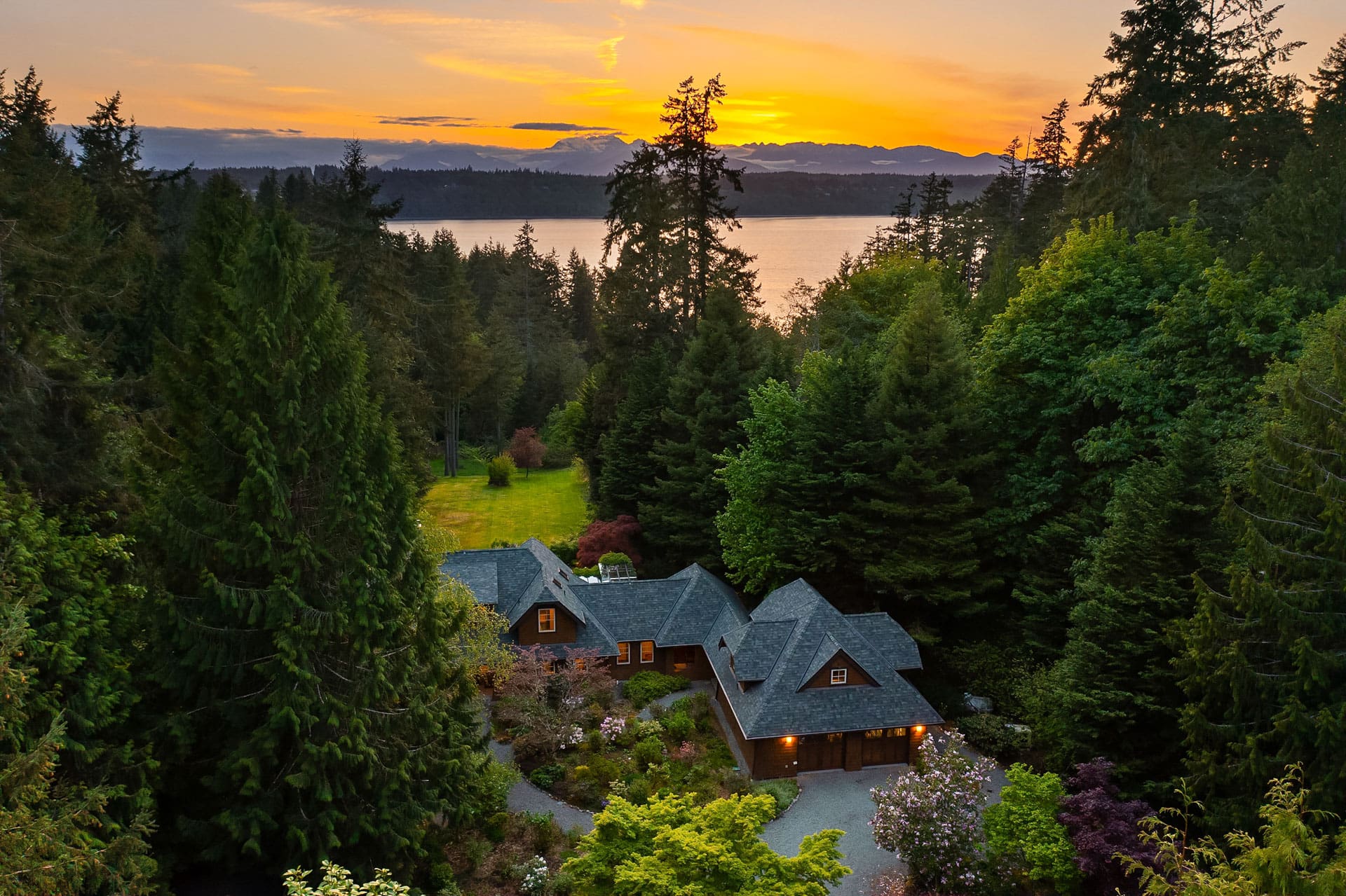 Elevated view of a charming house surrounded by lush evergreen trees, with a vibrant sunset over a distant lake. The sky is painted with warm hues of orange and yellow, reflecting across the serene water. The scene exudes tranquility and natural beauty.