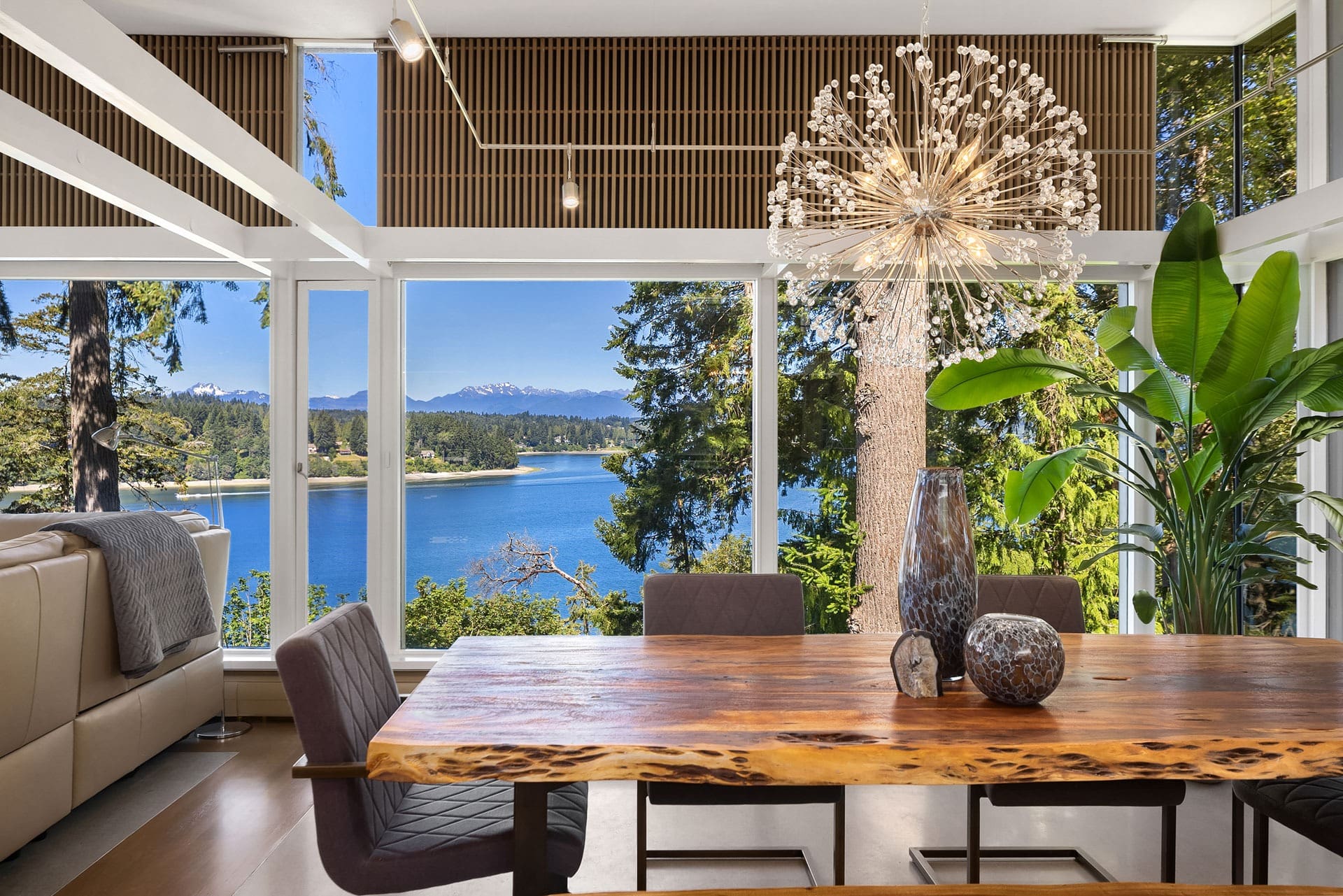 A dining area with a wooden table, modern chairs, and a decorative chandelier against a backdrop of large windows offering a stunning view of a lake, trees, and distant mountains. The room features natural decor elements and abundant natural light.