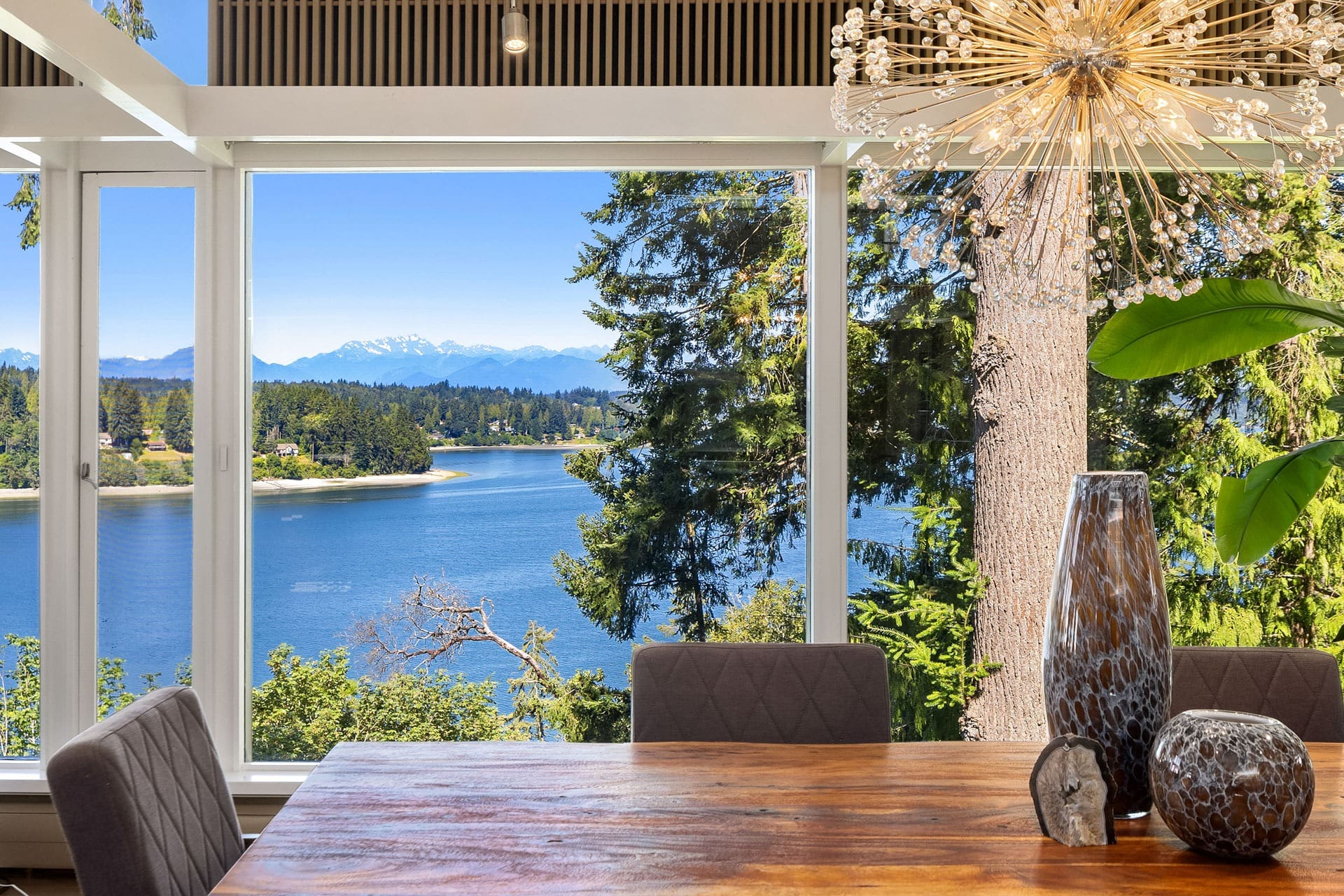 A dining table with decorative vases and a crystal chandelier in a room with large windows overlooks a scenic view of a lake, lush trees, and distant mountains under a clear blue sky.