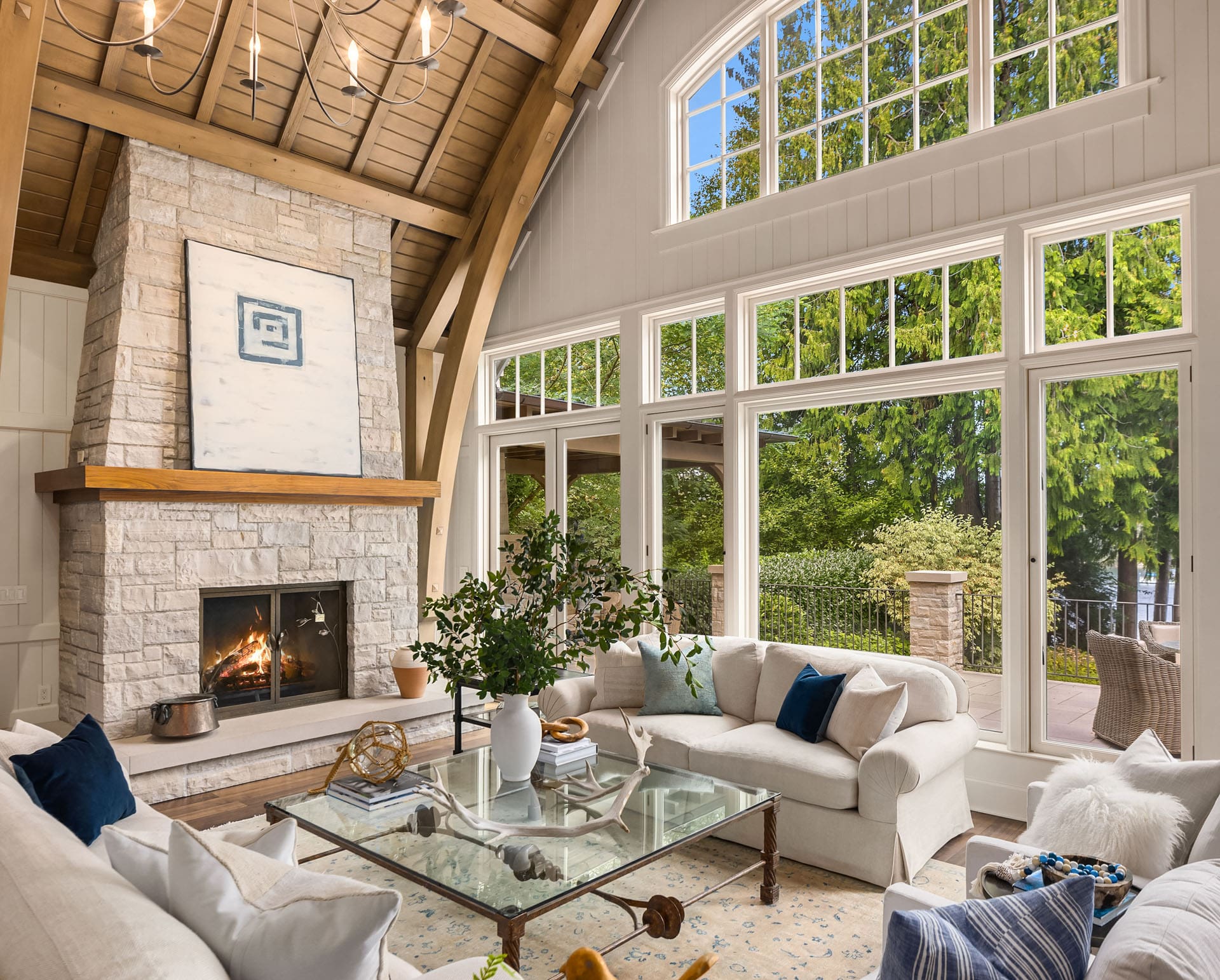 A cozy, elegantly designed living room features large windows, wooden beams, a stone fireplace, and white furnishings. A glass coffee table and greenery in a vase add a touch of nature. Outside, a lush green landscape is visible under a bright blue sky.