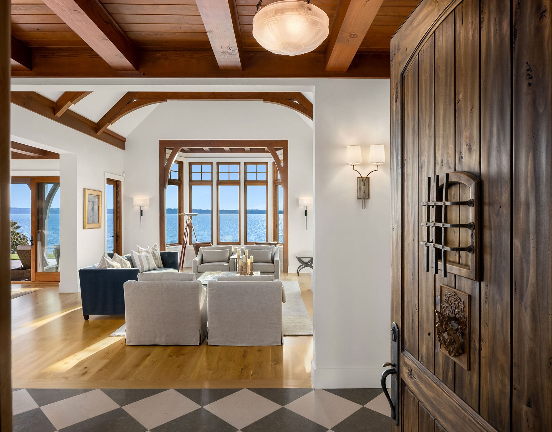 A cozy living room with wooden beams and large windows overlooks a serene ocean view. The room features comfortable seating with a mix of gray and dark blue sofas, wooden flooring, and minimalist decor. A wooden doorway leads into this inviting space.