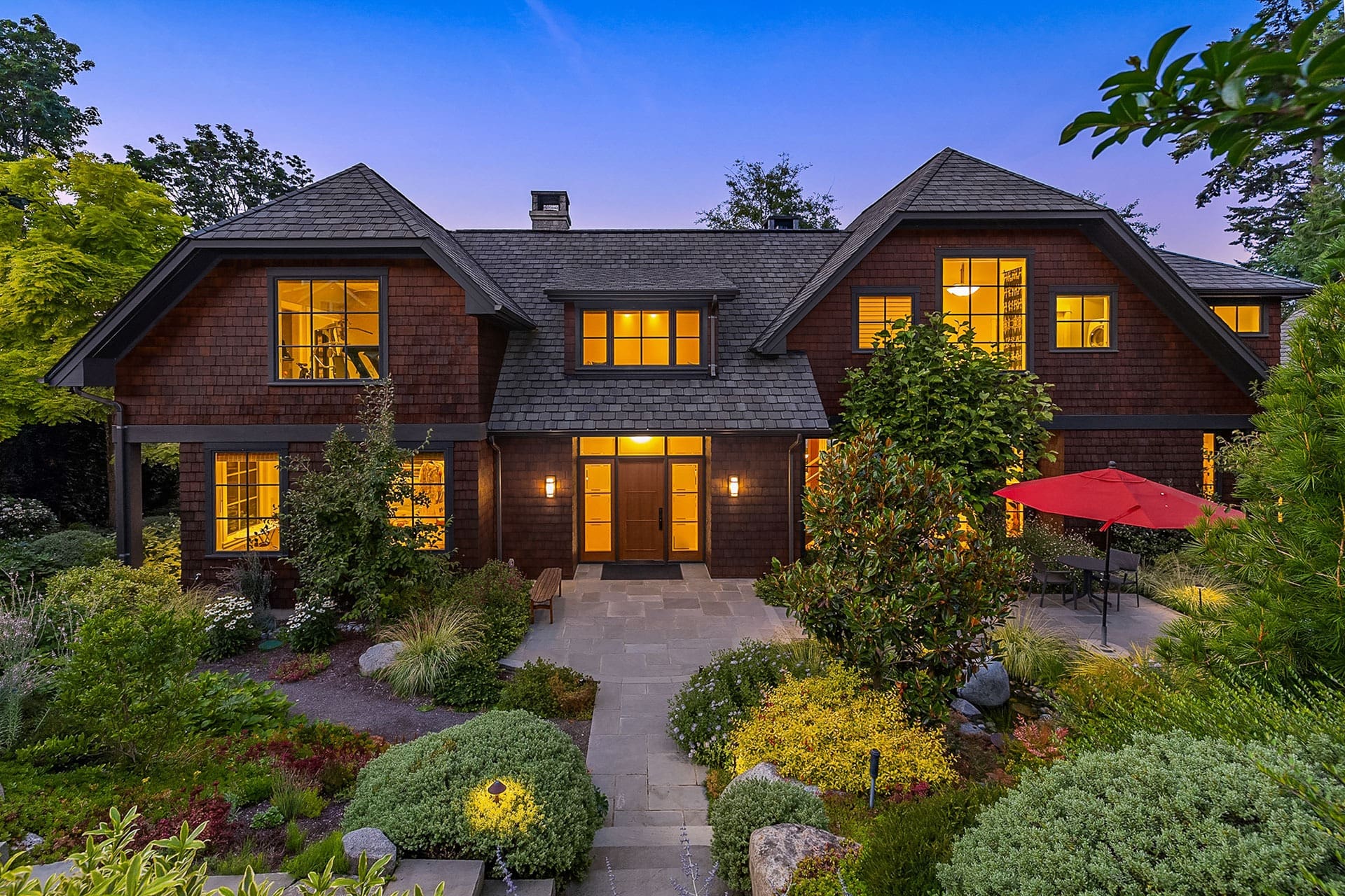 A two-story house with wooden siding and large, illuminated windows at dusk. The property includes a well-maintained garden with shrubs, trees, and a stone pathway. A patio area features a table and chairs under a red umbrella. The background shows trees and a clear sky.