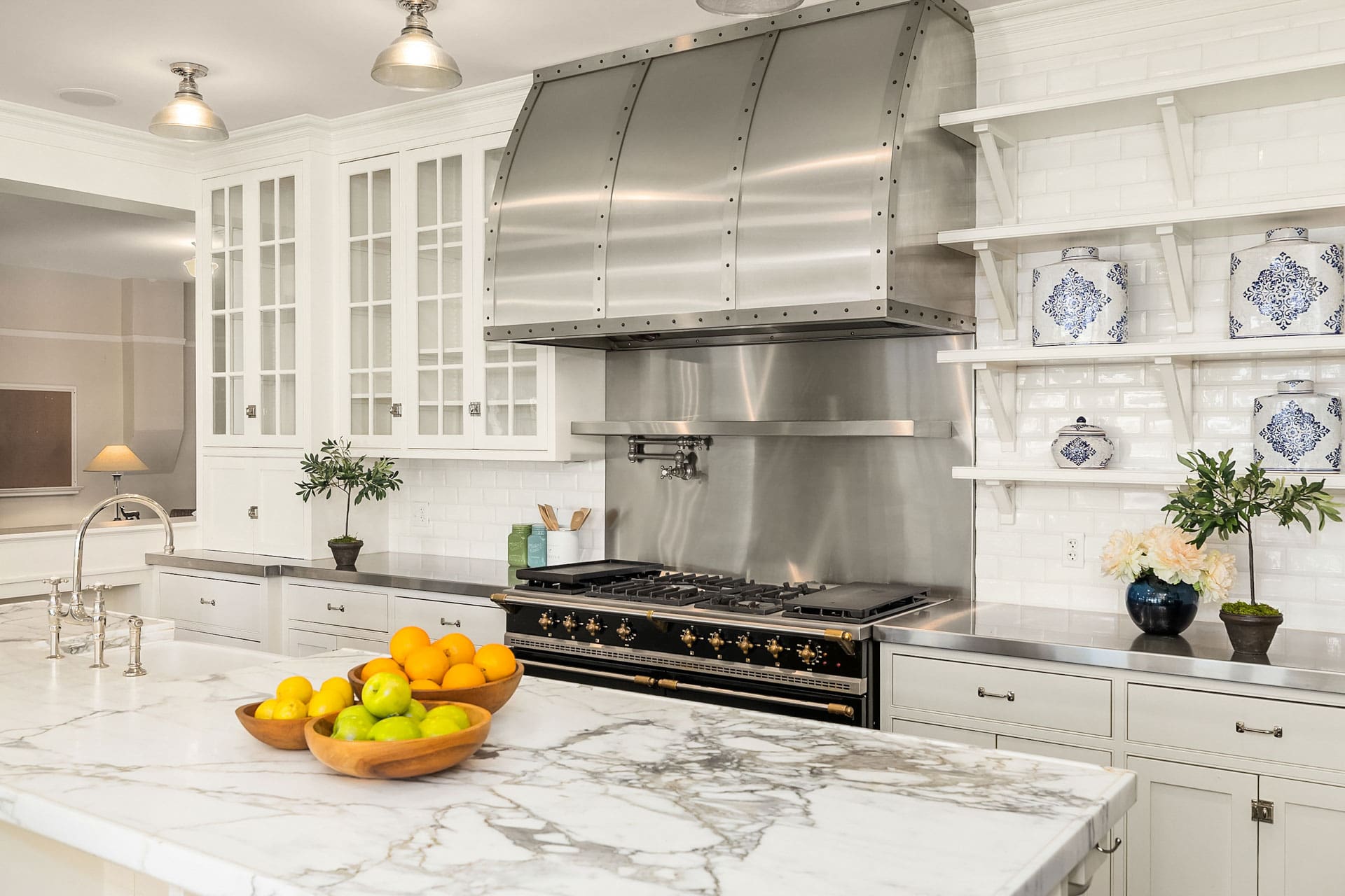 A modern kitchen with a large marble island featuring a wooden bowl of fruit. White cabinets with glass doors, stainless steel appliances, and a large hood over a black stove. Open shelves display blue and white decorative jars and green plants.