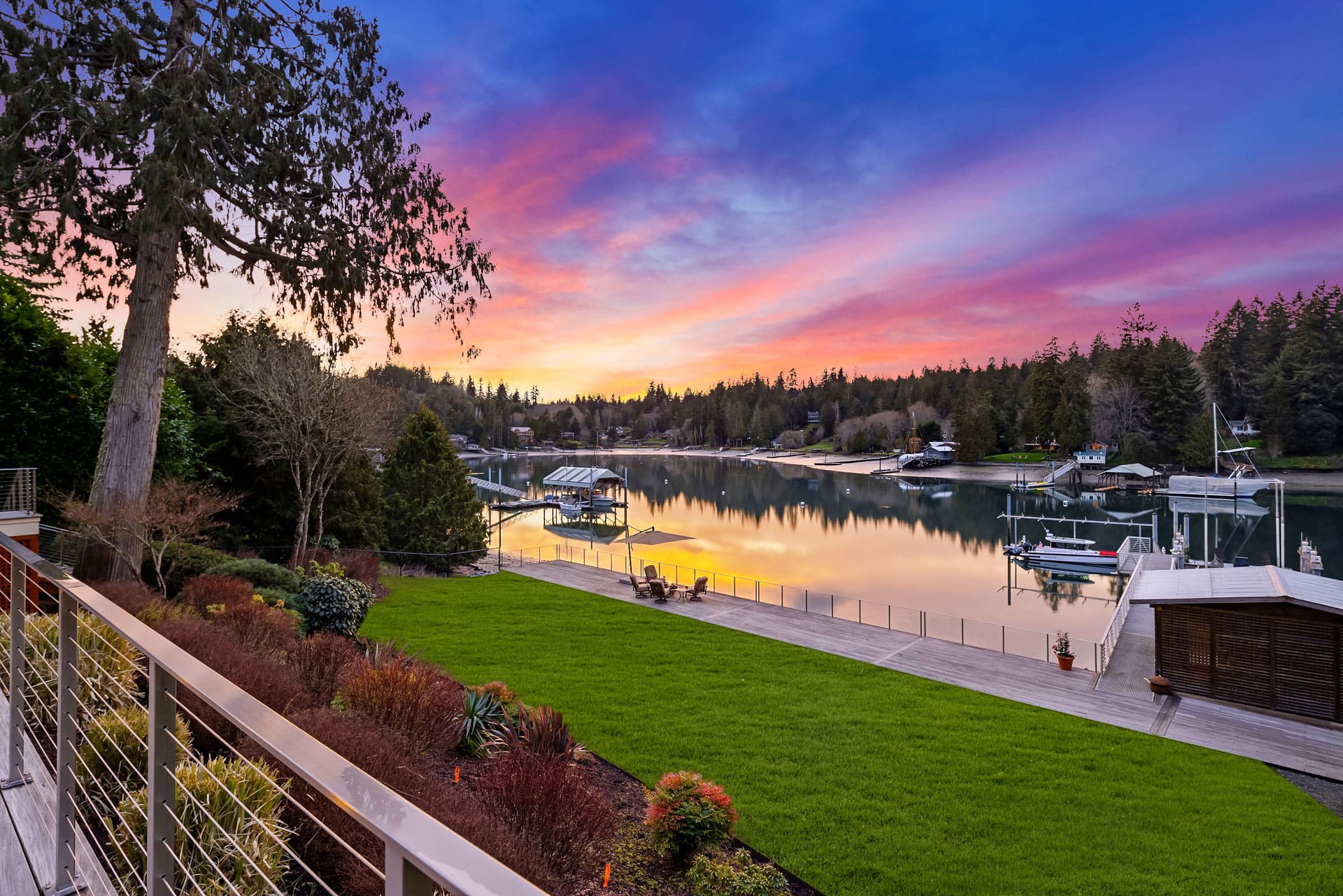 A tranquil lakeside scene at sunset featuring a well-maintained garden and a modern dock leading to the water. The sky is vibrant with pink, purple, and orange hues, reflecting on the calm water. Surrounded by tall trees, the area feels serene and picturesque. A charming, dark brown cottage with illuminated windows stands amidst a landscaped lawn at dusk. A small tree with a spotlight and an outdoor seating area enhance the cozy atmosphere. The clear sky overhead features a crescent moon, and tall trees frame the background. Twilight shot.