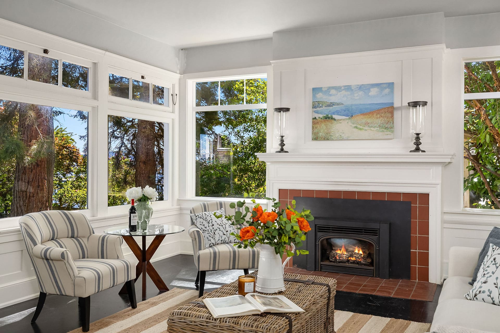 A cozy living room features a fireplace with a painting above it, flanked by two large windows showing scenic outdoor views. Two striped armchairs face each other with a small round table in between, topped with flowers. A wicker coffee table holds a vase of orange flowers and an open book.