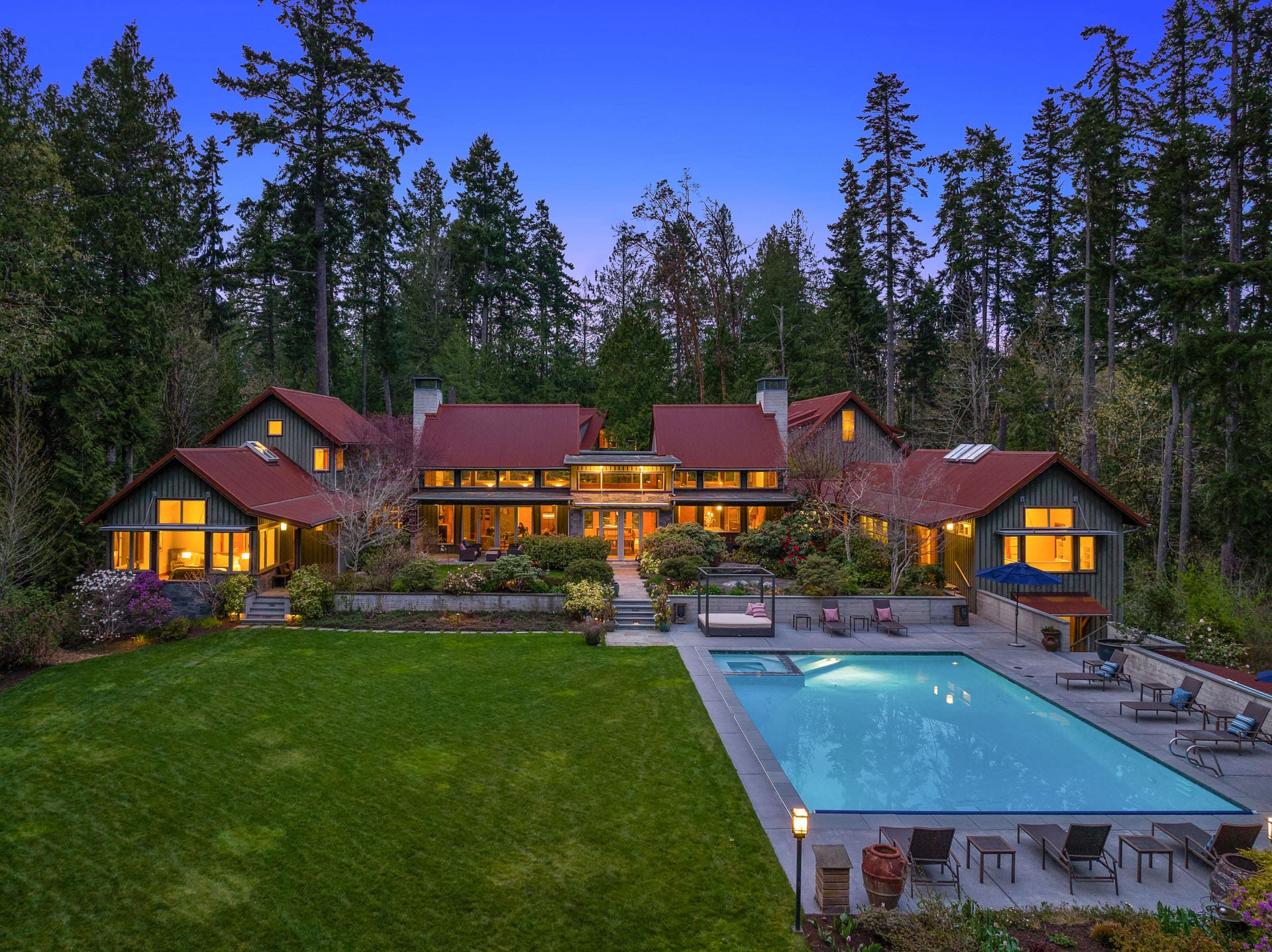 A large, luxurious house with red roofs, surrounded by tall trees. The well-lit home features expansive windows and a sprawling green lawn. In the backyard, there's a well-maintained swimming pool with patio seating, blending naturally with the forested landscape.