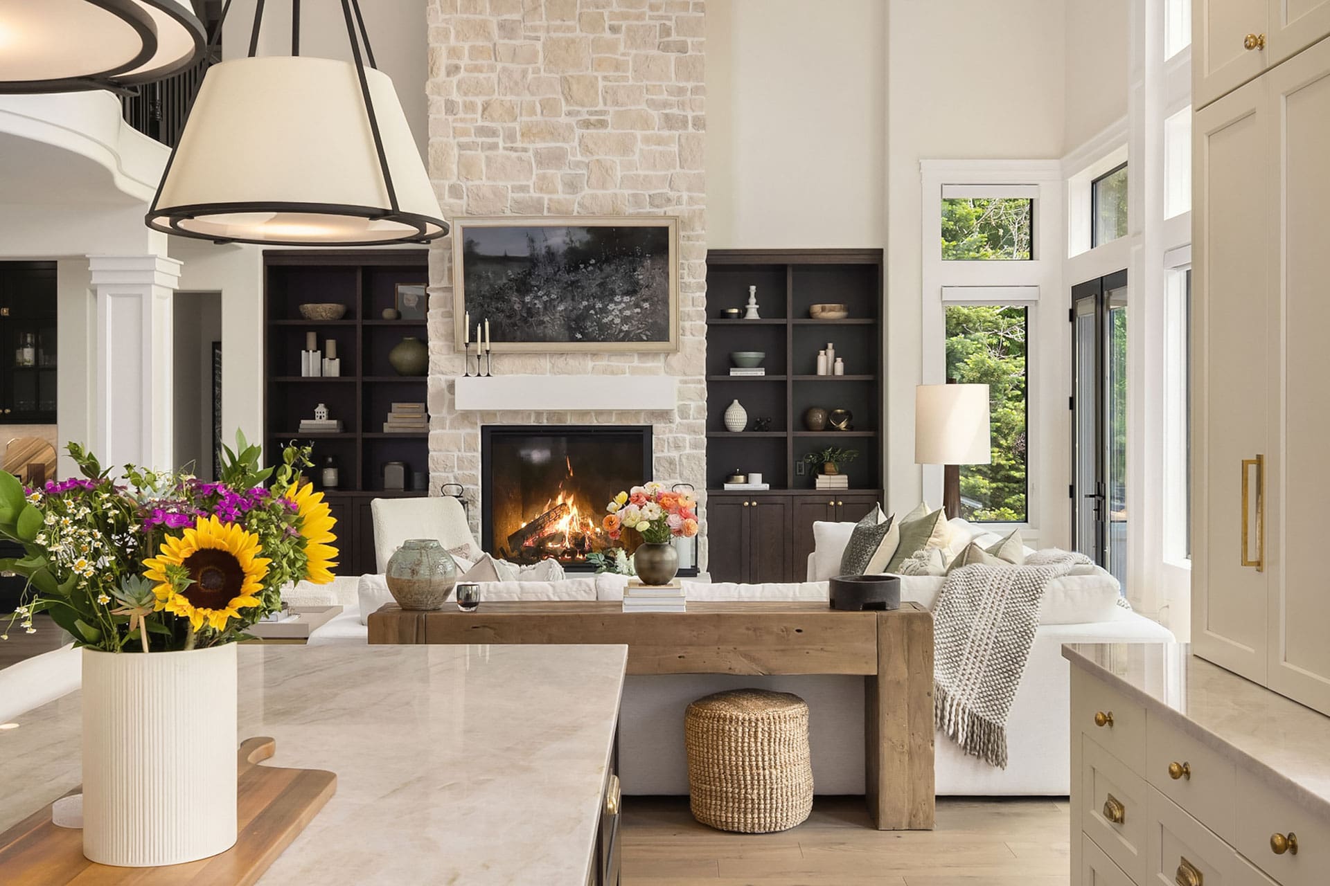 Modern open-plan living room with a high ceiling, stone fireplace, and built-in shelves. White sofa with colorful cushions and fresh flowers on a wooden coffee table. Large windows provide natural light. Kitchen island with flowers in the foreground.