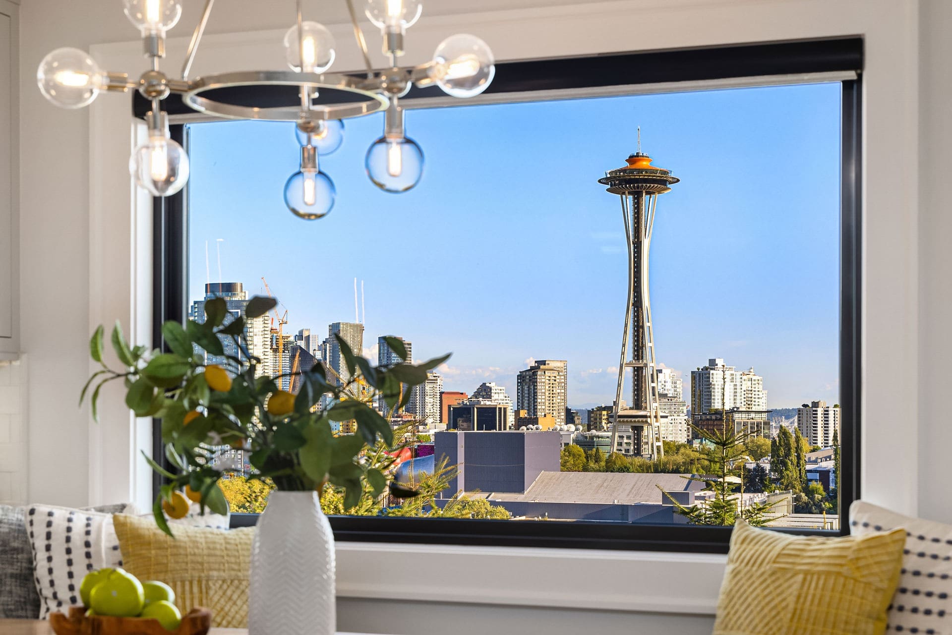 A bright, modern room features a large window with a view of the Seattle Space Needle and city skyline. The window is framed by a light fixture with hanging bulbs. A vase with green foliage and lemons sits on a table next to a yellow and white cushion.