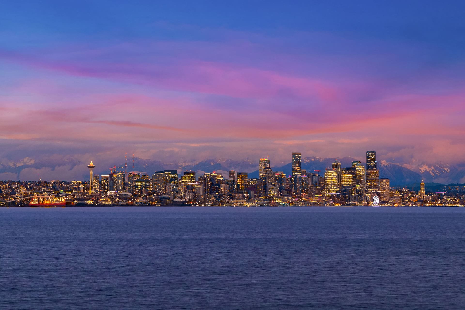 A panoramic view of a city skyline at dusk, with tall, illuminated buildings against a sky transitioning through shades of blue and pink. In the foreground is a calm body of water, and a prominent Ferris wheel and distinctively shaped tower are visible in the cityscape.