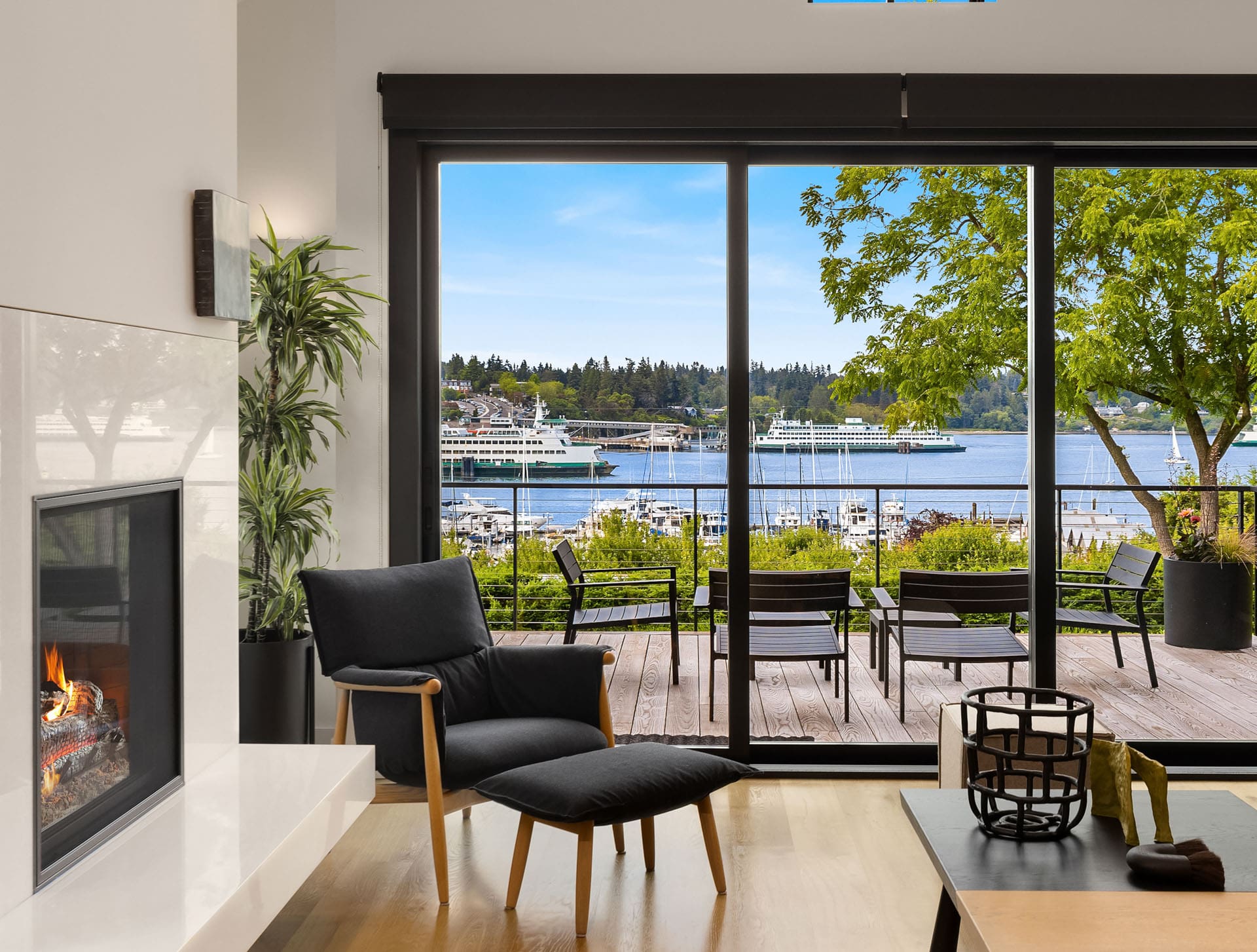 A modern living room features a sleek fireplace, an armchair with an ottoman, and a minimalist coffee table. Floor-to-ceiling glass doors open to a wooden patio with chairs, overlooking a marina with docked boats and a scenic view of a tree-lined waterfront.