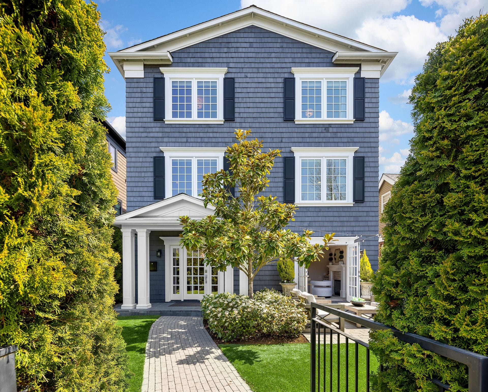 A three-story house with a gray facade, white trim, and blue shutters. The entrance is framed by a white portico, and the front yard features a neatly manicured lawn, shrubs, and a brick walkway. Tall, green trees stand on both sides of the property.