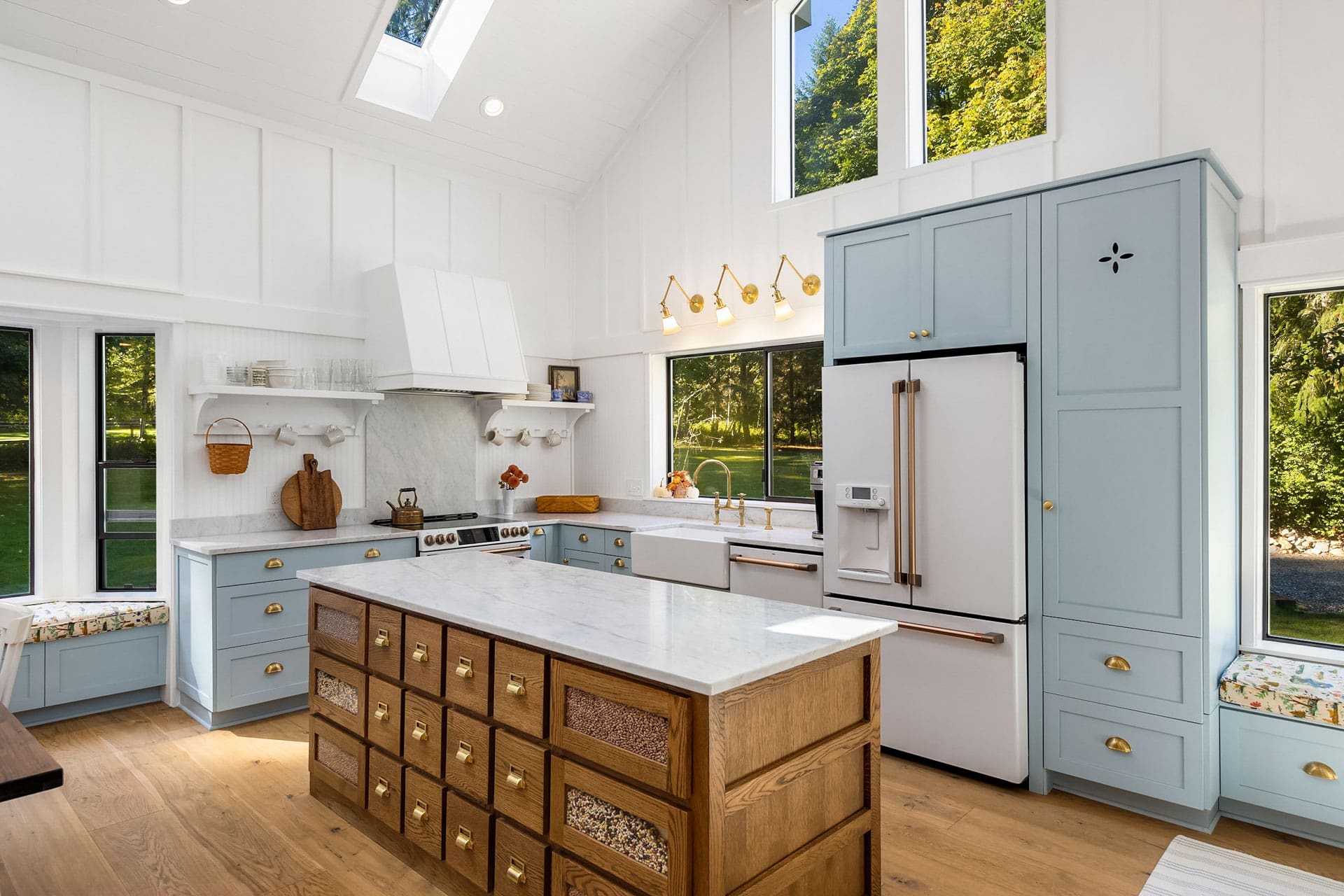 A bright, modern kitchen with white paneled walls and tall ceilings featuring skylights. It has blue cabinetry, a wooden island with marble countertop, built-in appliances including a white fridge, gold accents, large windows, and wooden flooring.