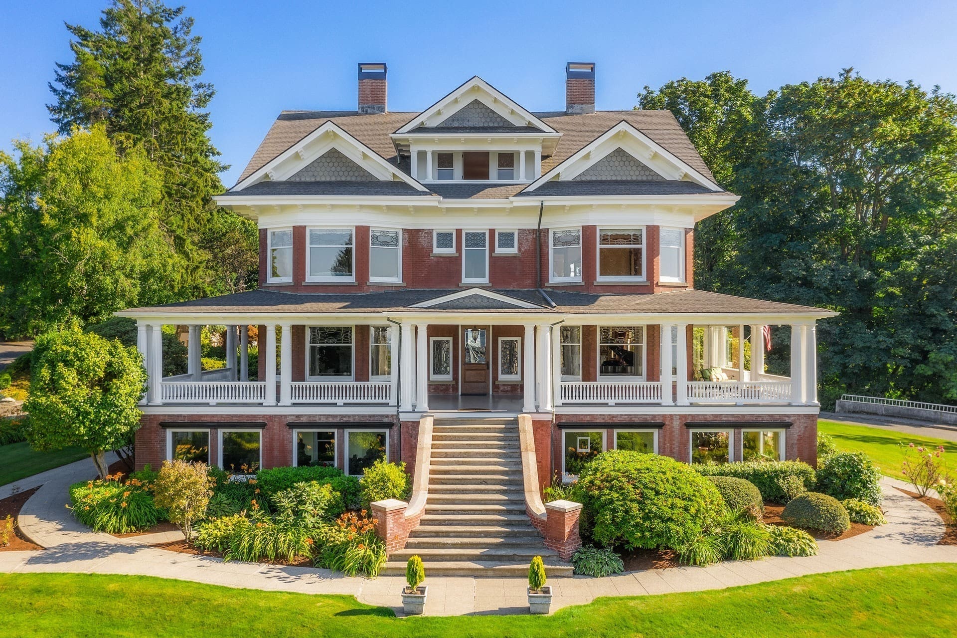 A large, two-story brick house with a wraparound porch and white columns, set in a lush garden. The house features a wide staircase leading up to the entrance, large windows, and a well-maintained lawn with bushes and trees. The sky is clear and blue.