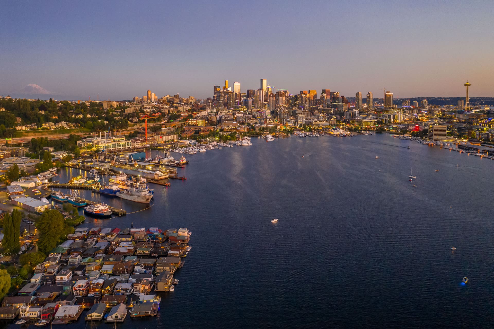 Aerial view of Seattle's Lake Union during sunset, with boats docked along the waterfront. The city skyline, including the Space Needle, is visible in the background. The water reflects the warm hues of the setting sun, and Mount Rainier is faintly visible on the horizon.