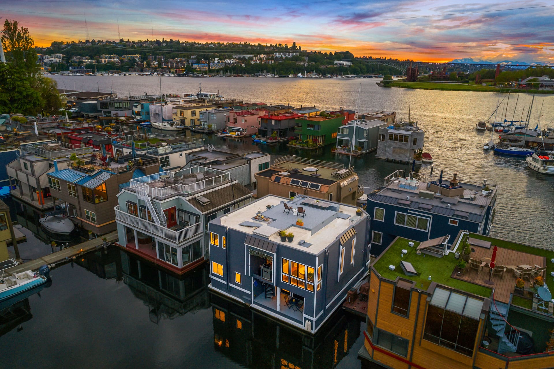 Aerial view of colorful houseboats on a tranquil lake at sunset. The sky is painted with shades of orange, pink, and blue. Trees and buildings are seen in the distance, and several boats are docked along the shore. The houseboats have varying designs and rooftop decks.