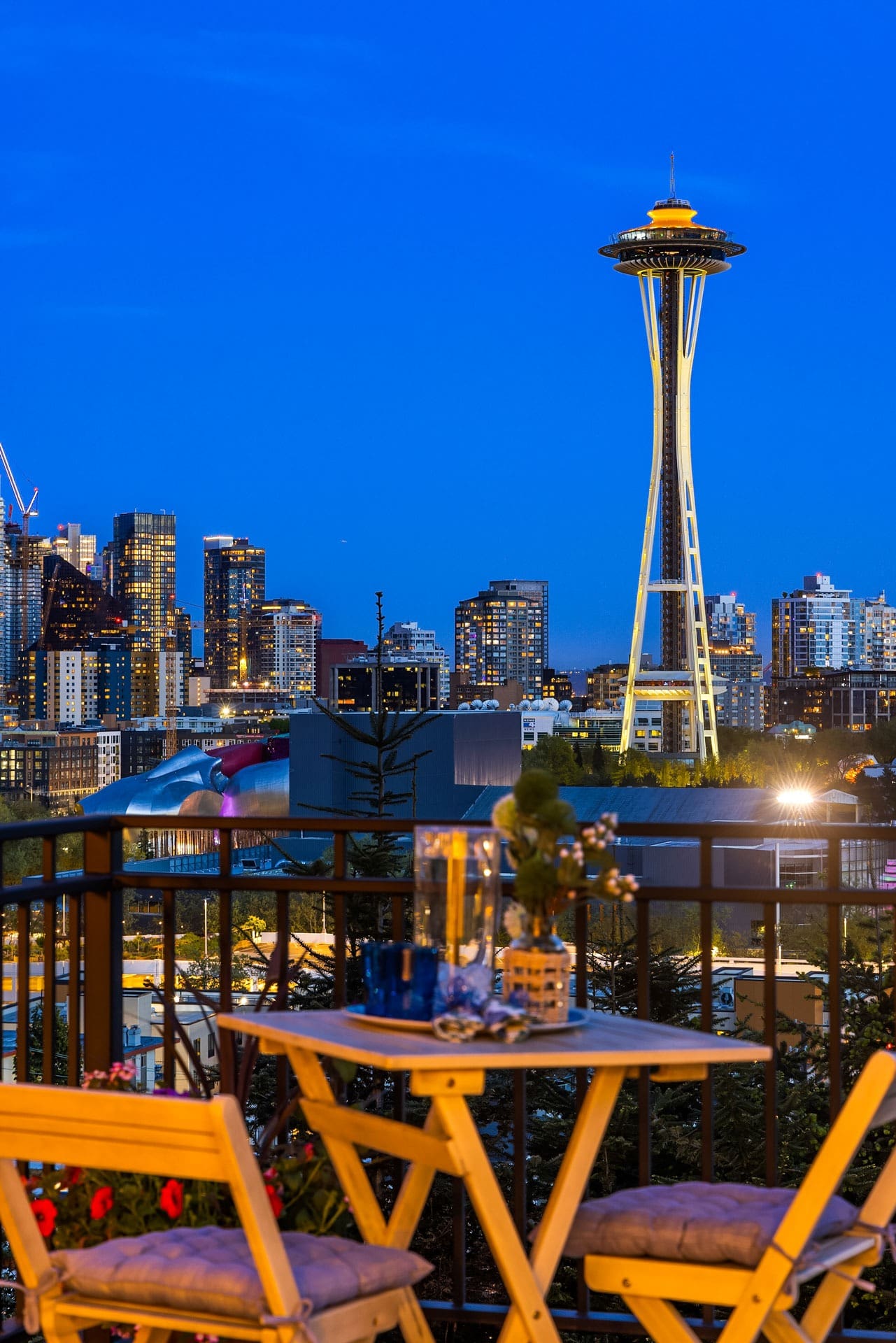 Cozy outdoor seating area with wooden chairs and a small table set on a balcony overlooking a night cityscape. Iconic Space Needle lit up against a deep blue sky, surrounded by illuminated modern buildings. Potted plants add a touch of greenery.