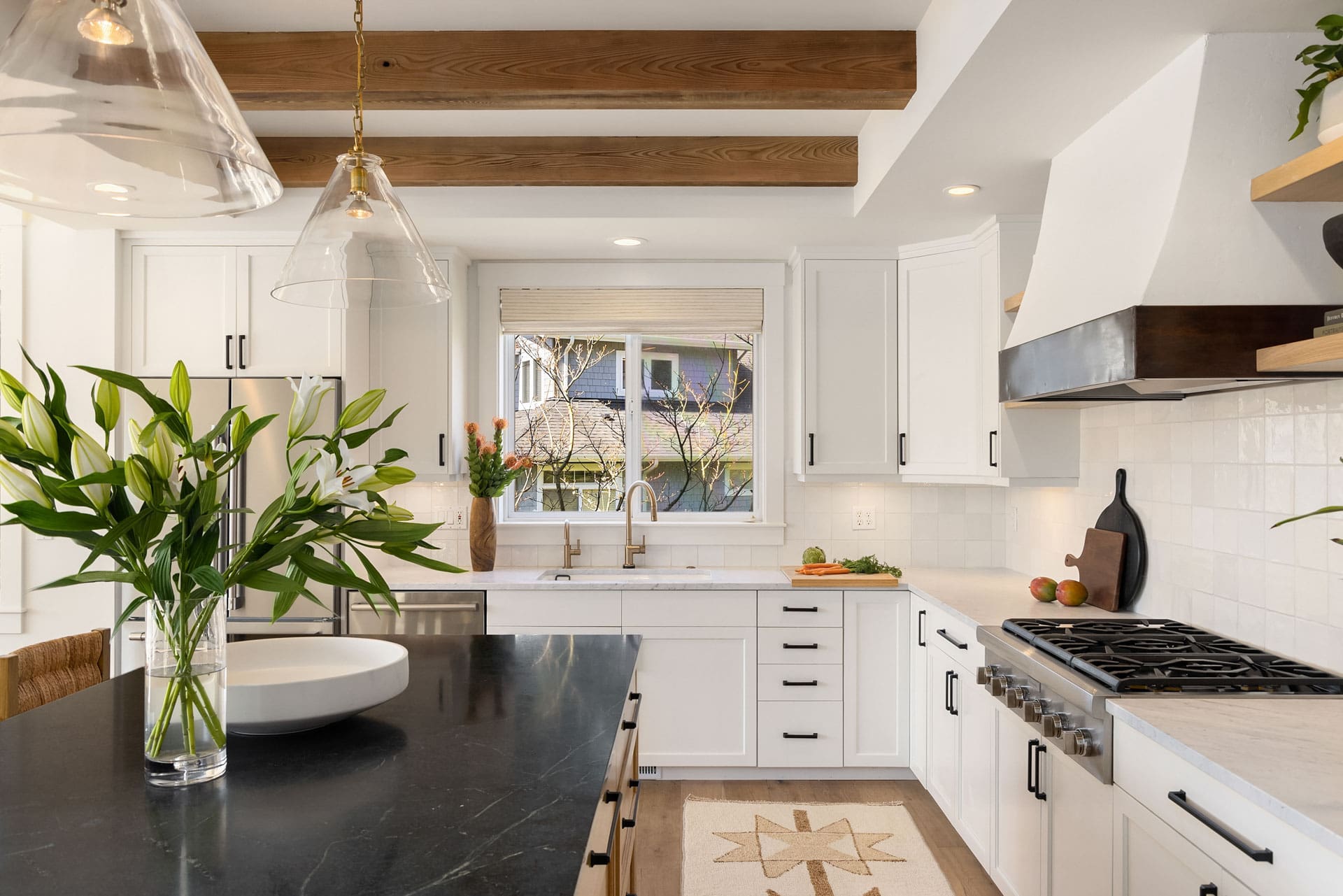 A modern, bright kitchen with white cabinets, a black countertop island, and wooden ceiling beams. The island features a vase with white lilies and pendant lights above. Natural light streams through a window above the sink, illuminating plants and decor.