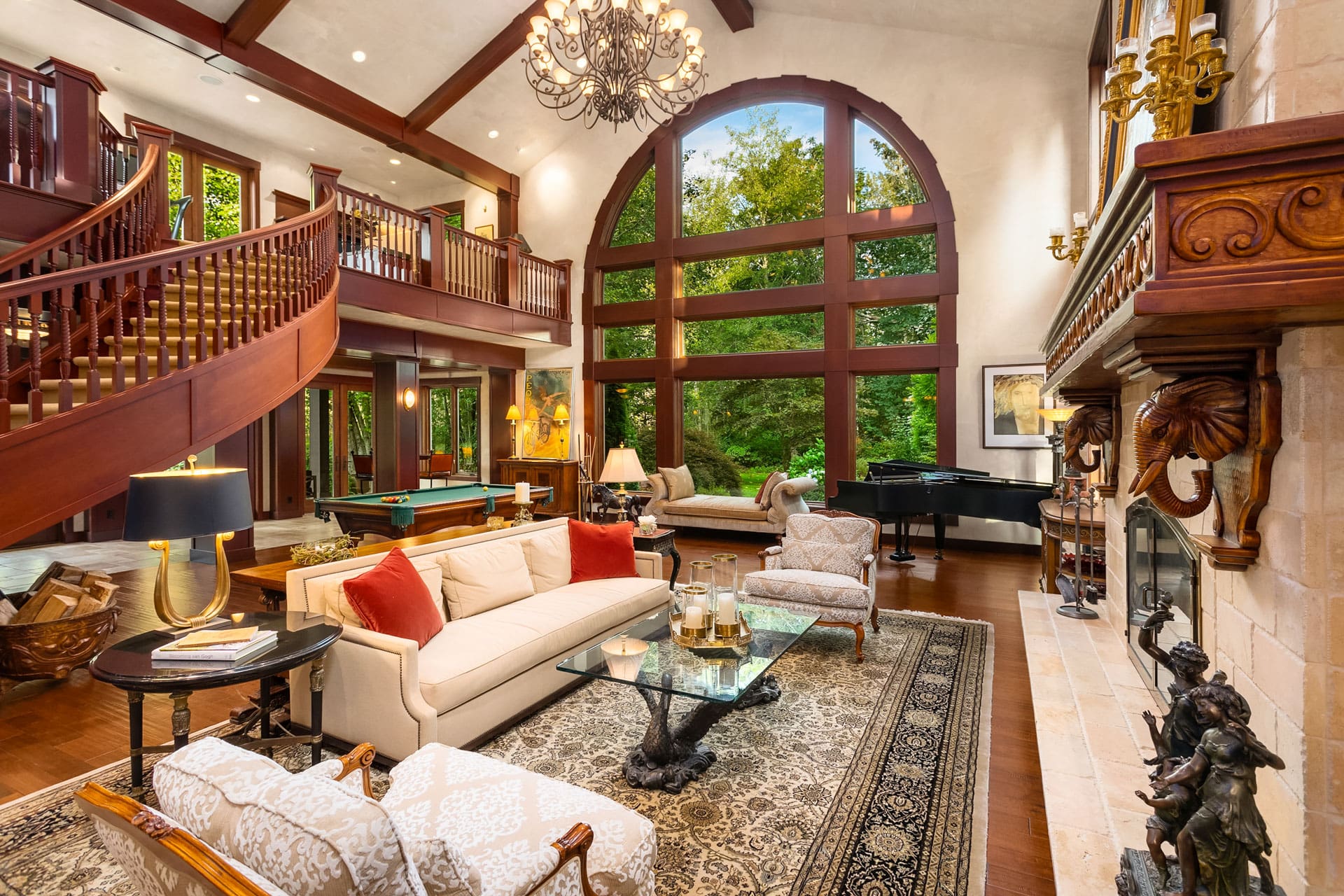 A spacious and elegant living room features a curved staircase, high ceilings with wooden beams, large arched windows offering a view of lush greenery, a chandelier, a fireplace, comfortable seating, a grand piano, and a mix of traditional and modern decor.