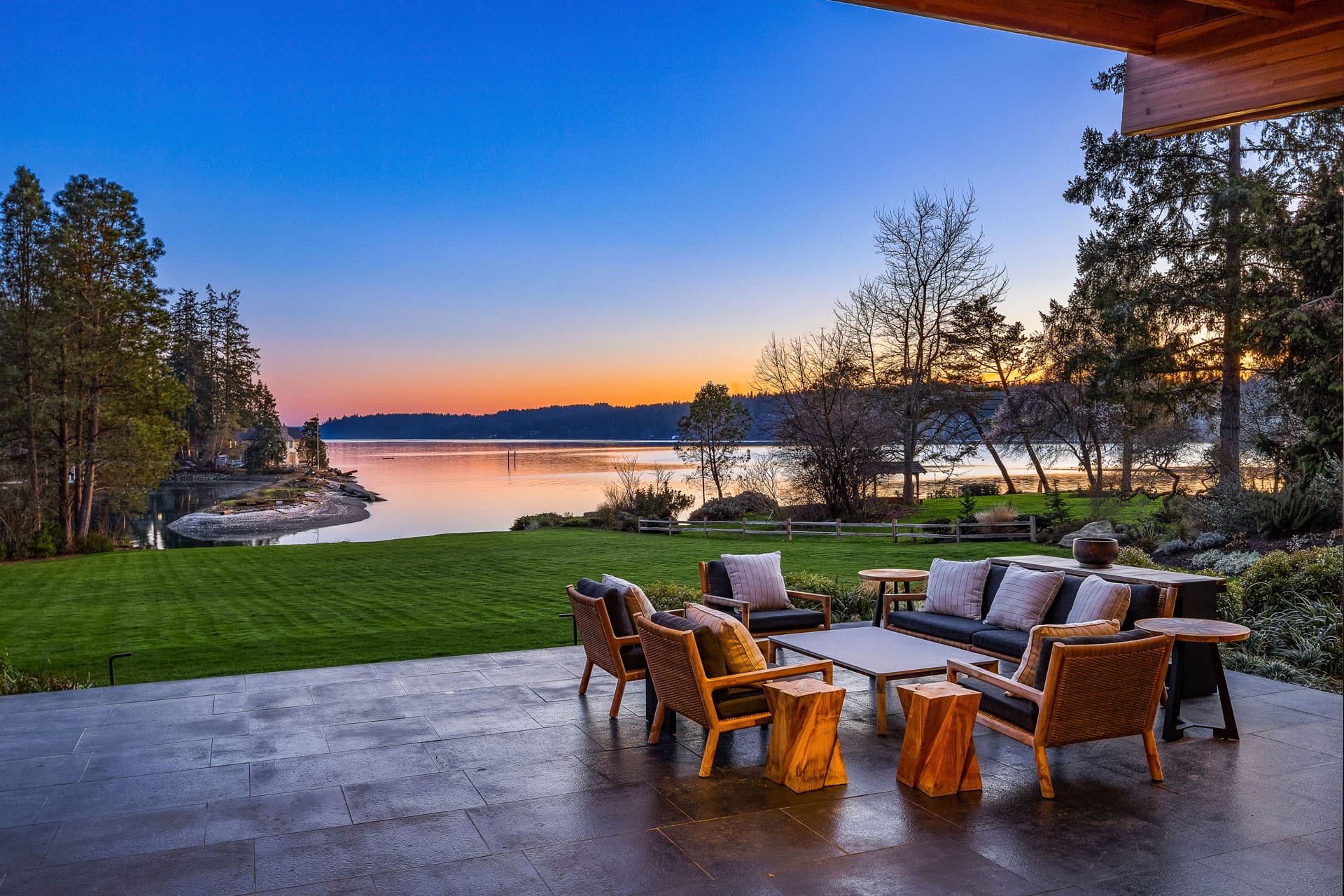 A serene lakeside view at sunset features a cozy outdoor seating area on a stone patio. Wooden chairs with cushions surround two small wooden tables, set against a backdrop of a lush green lawn and calm water reflecting the twilight colors. Trees line the shore.