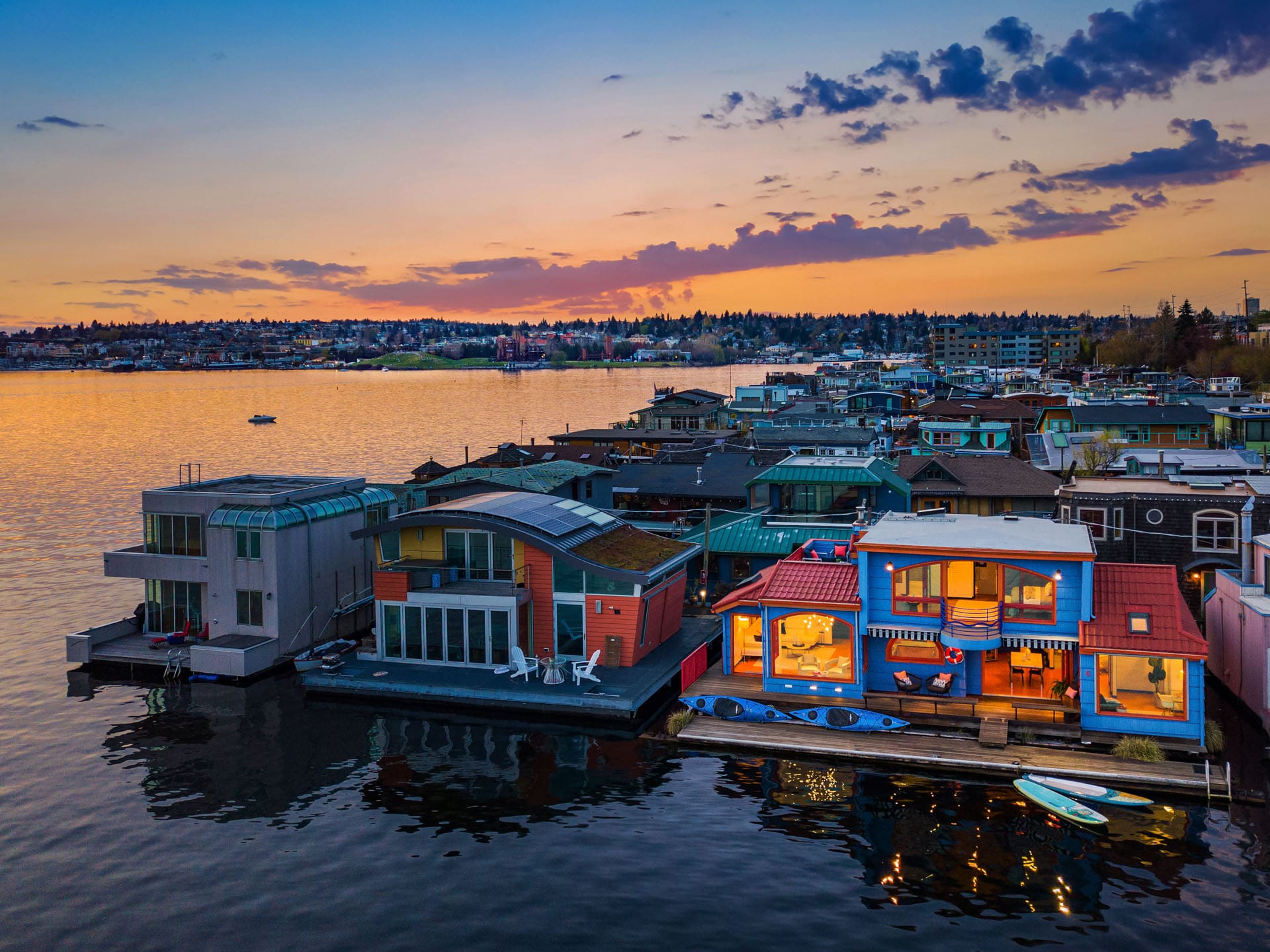 Sunset view of a picturesque floating house community on a calm lake. The houses, adorned with vibrant colors and large windows, reflect on the water. The sky is painted in warm hues of orange, pink, and blue as the day transitions to evening.