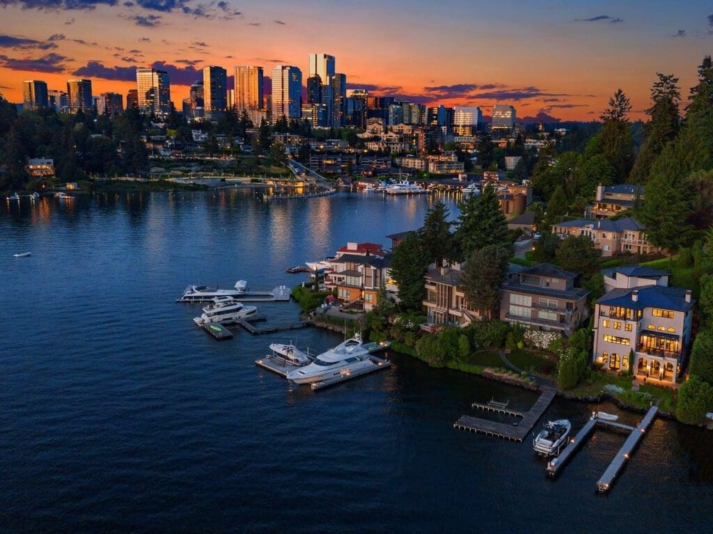 Aerial view of a suburban waterfront neighborhood during sunset. Luxury houses with private docks and boats line the shore. In the background, a city skyline is illuminated against a colorful dusk sky, with reflections visible on the water surface.