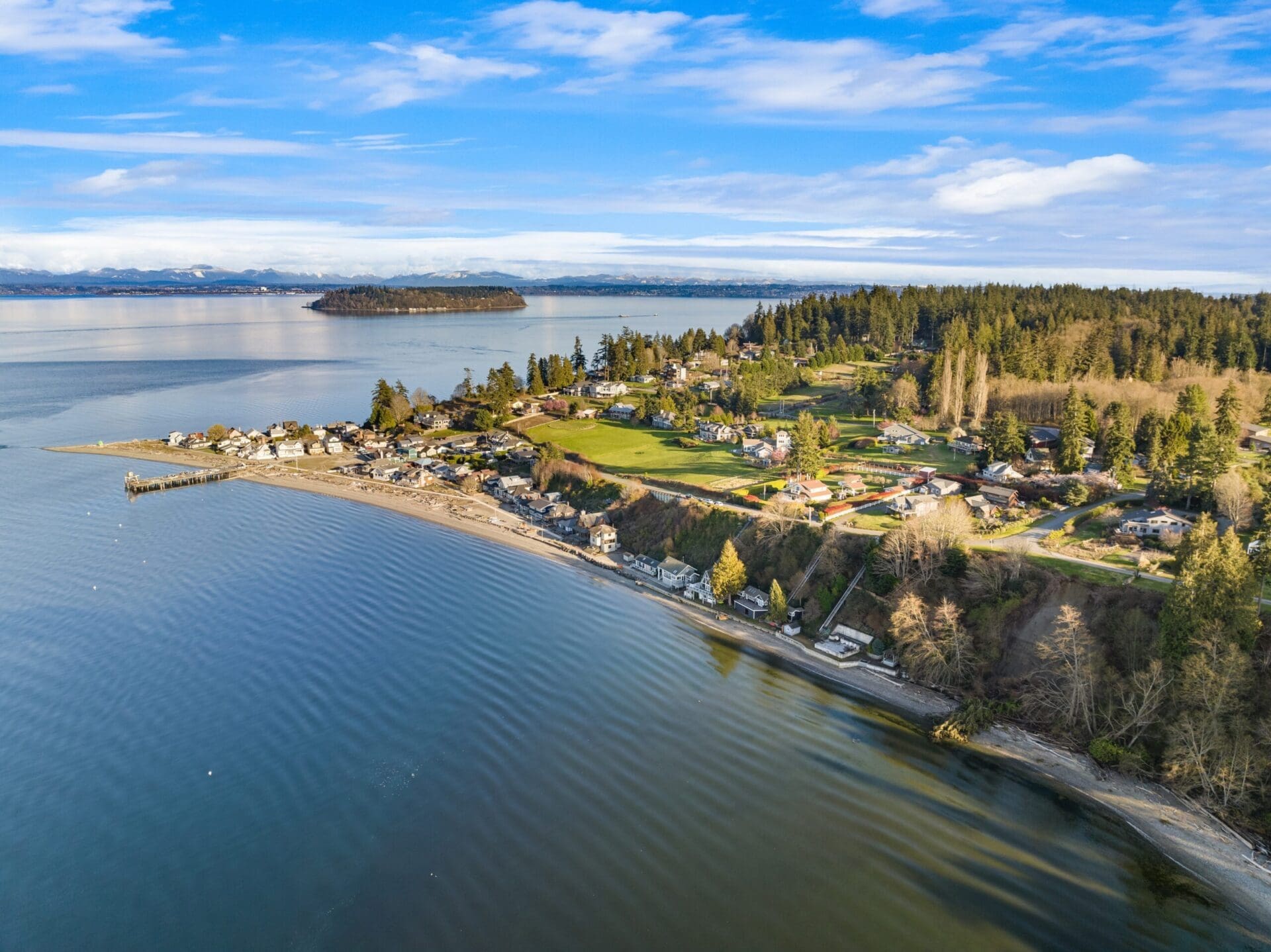 Aerial view of a coastal town featuring houses along a sandy shoreline, surrounded by evergreen trees. The calm ocean stretches out to the horizon with an island visible in the distance. The sky is clear with scattered clouds, and the scene is brightly lit by the sun.