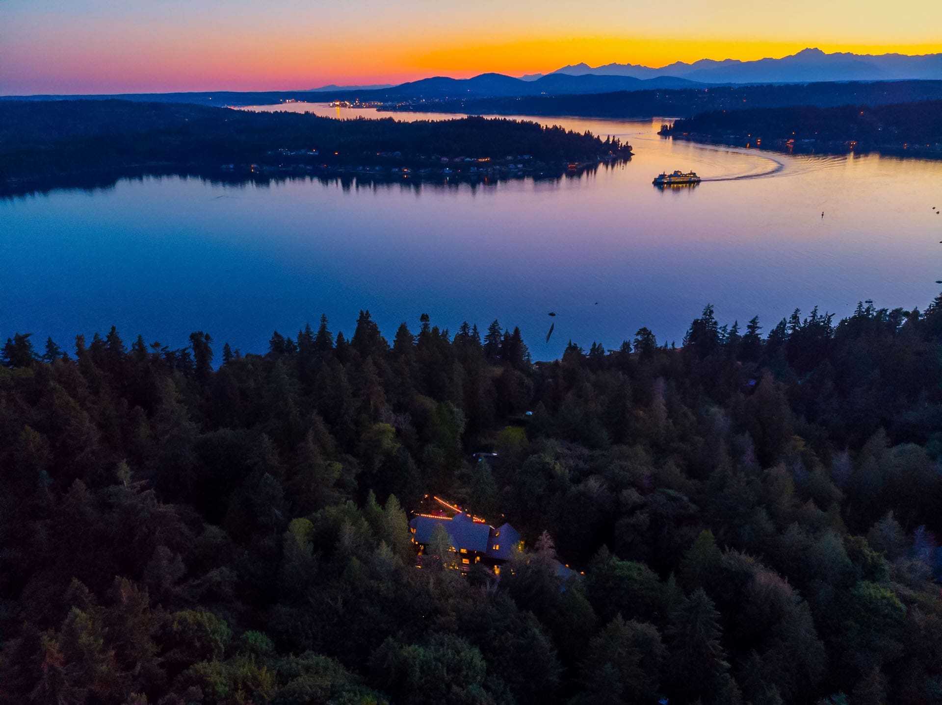 Aerial view at dusk of a tranquil lake surrounded by dense forest and distant mountains. A ferry is visible on the lake, and a house with glowing lights is nestled amongst the trees in the foreground, creating a serene and picturesque scene.