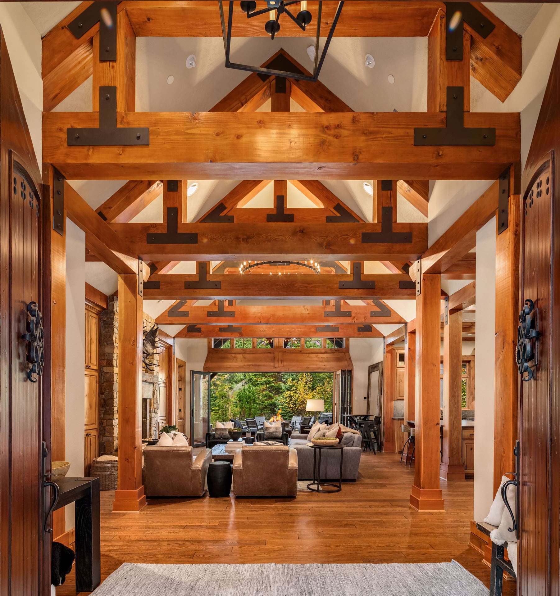 A spacious, rustic-style living room with high exposed wooden beams, large windows, and a mix of beige and brown furniture. The room includes sofas, armchairs, and a coffee table, all arranged in front of a wide doorway leading to an outdoor view.