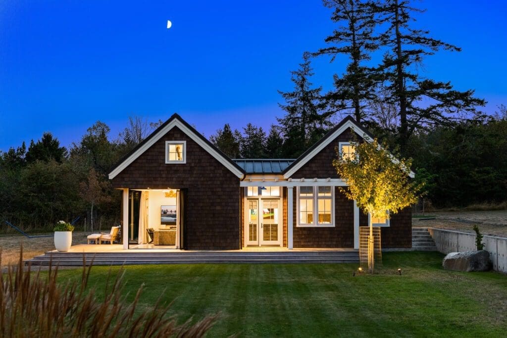 A cozy, well-lit house with dark wooden exterior walls and a spacious lawn is shown at dusk. The cottage-style home features large windows, a covered porch, and is surrounded by trees. The moon is visible in the twilight sky, adding to the serene ambiance.