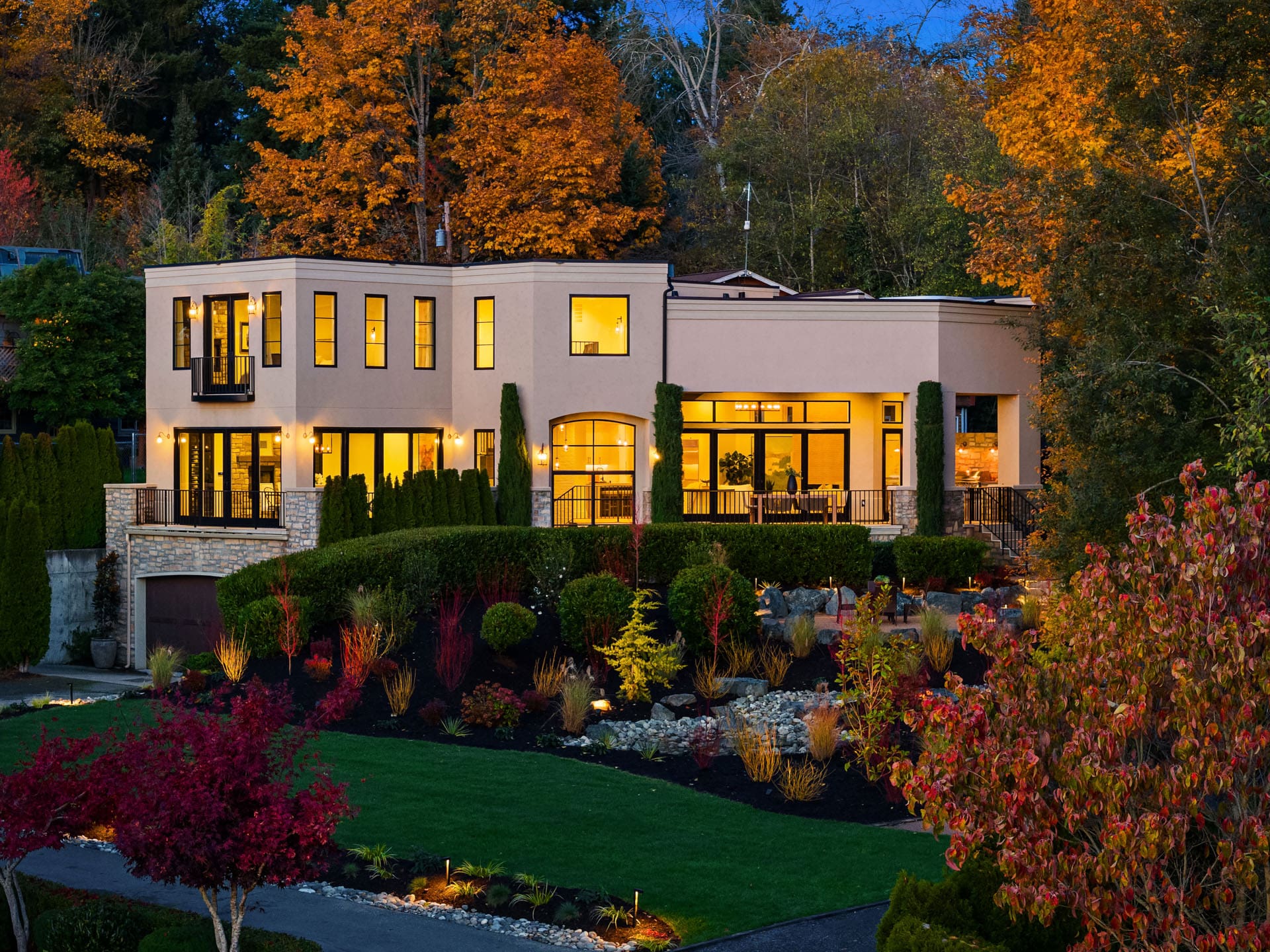 A modern two-story house with large windows, surrounded by lush landscaping and autumn-colored trees. The home features a mix of stucco and stone exterior, with a spacious lawn and well-lit pathways leading to the entrance. The scene is set during twilight.